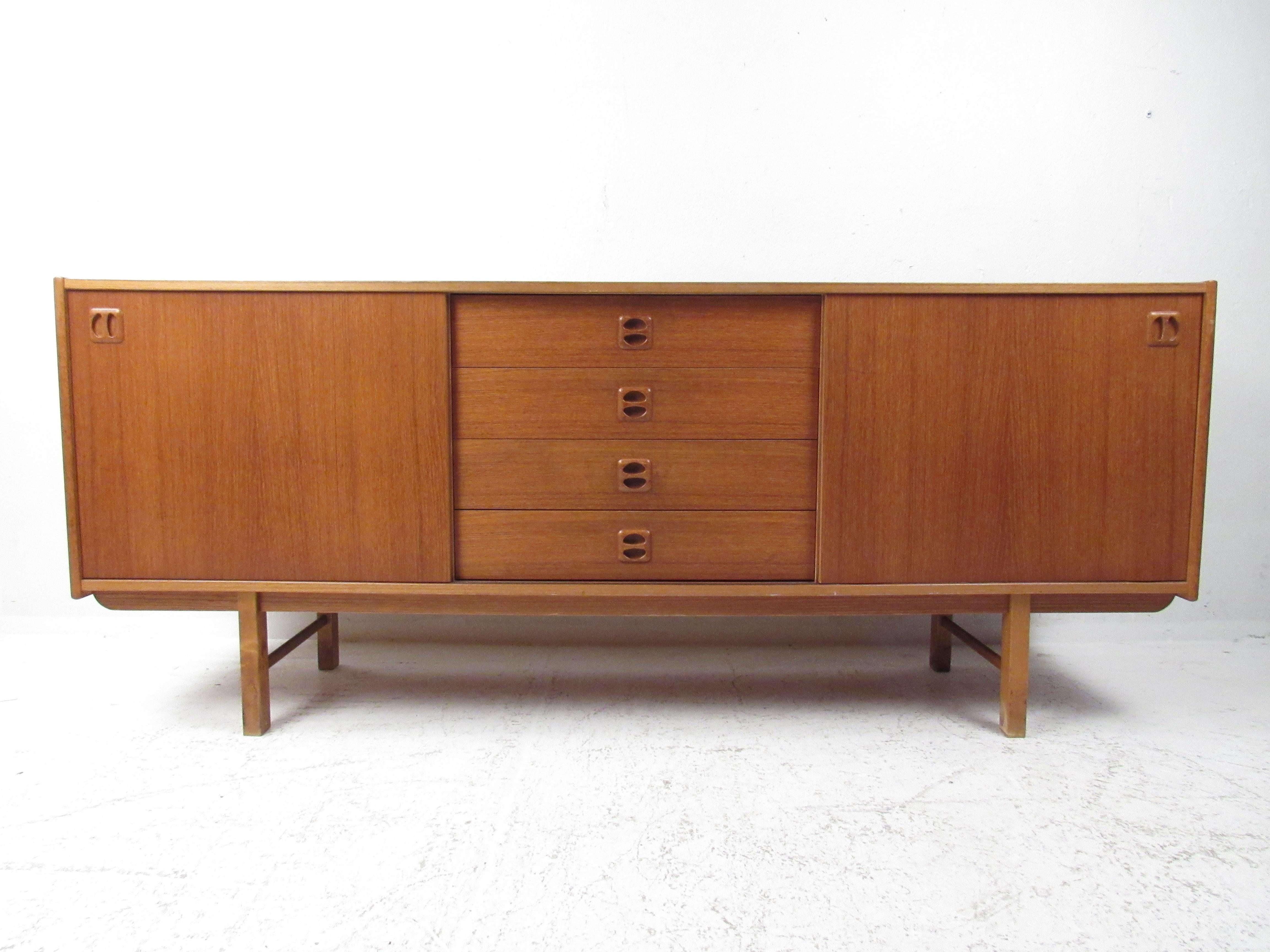 This stunning vintage modern sideboard offers plenty of room for storage within its four hefty drawers and two large compartments with shelves. Danish Modern design with sculpted recessed pulls, tapered legs, and a rich teak finish. A straight line