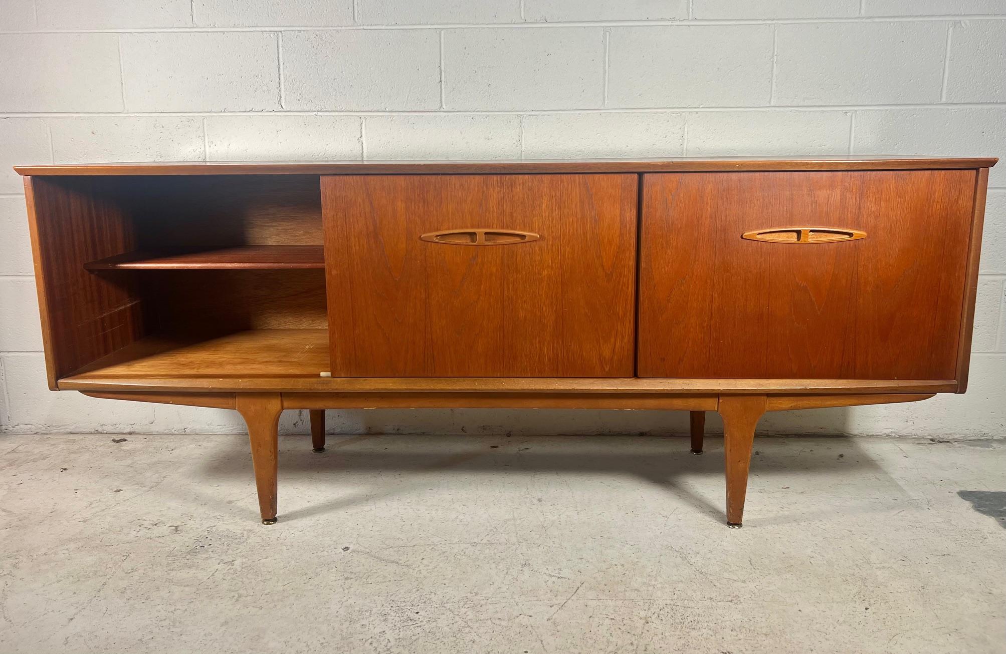 1960's teak veneer credenza with stained beech legs and handles by Jentique. Made in England. Original label in top drawer.

Three drawers in the middle flanked by sliding door cabinets with a shelf on either side. Top drawer has the original green