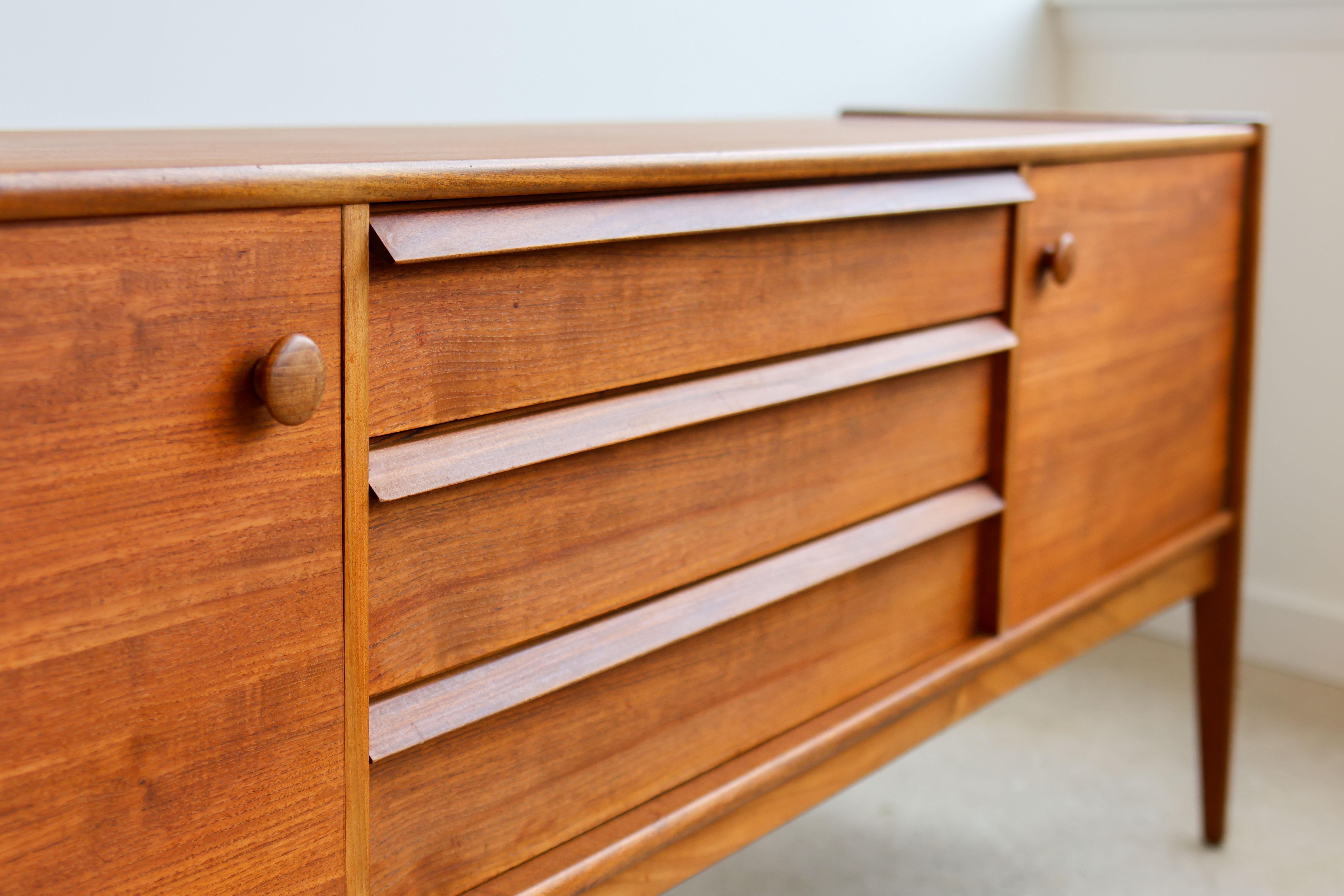 Mid-Century Modern teakwood credenza. 
Made by Younger Furniture Co, Ltd. 
Two cabinets:  1 with capabilities to hold records, 1 with built-in shelf.
Three dovetailed drawers; top drawer holds dividers.
Rarely seen finished back.
Excellent vintage