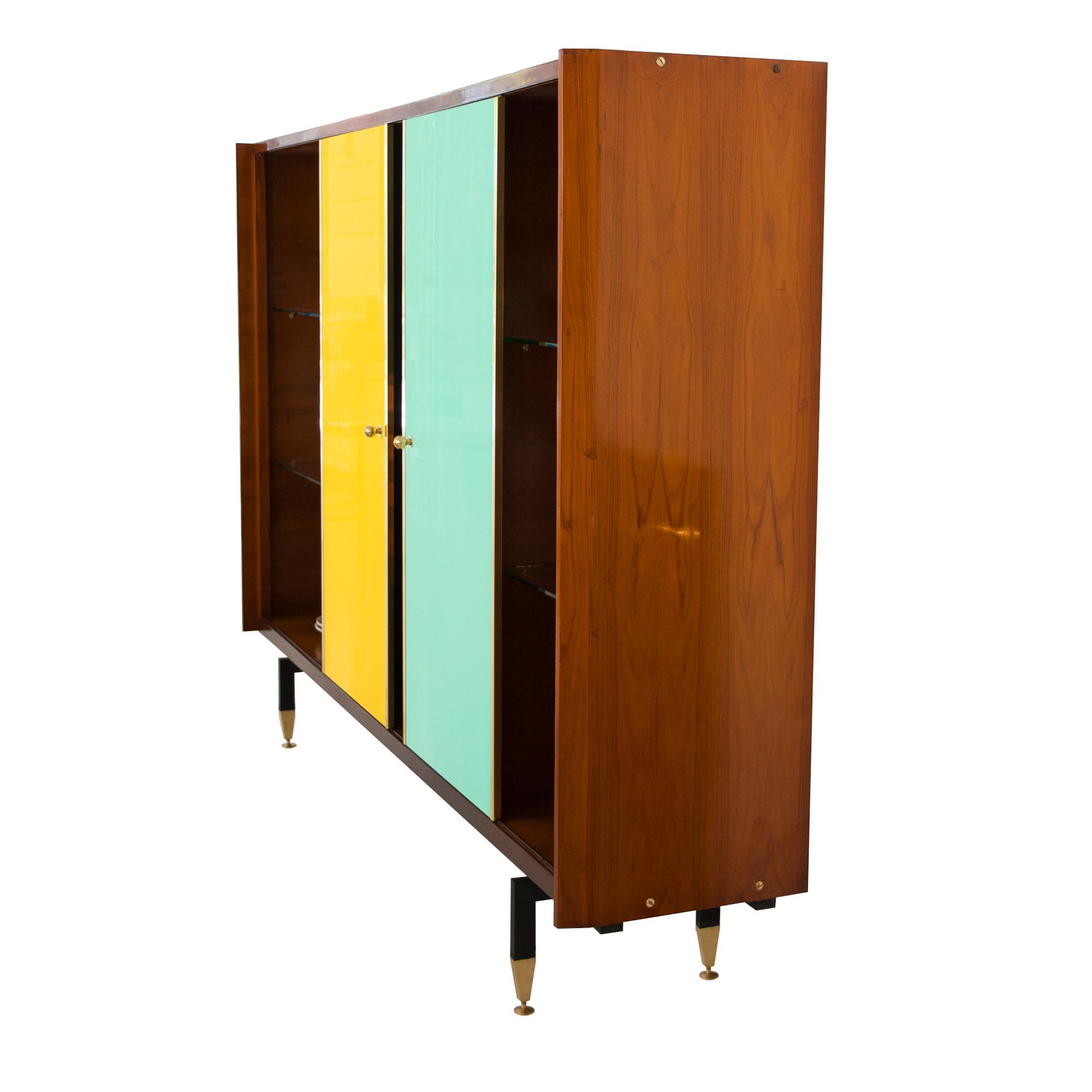 Sideboard made of teak with colored glass sliding doors with brass finishes. Composed of 3 sections, 2 sides with tempered glass shelves and a central one with two decorative holes and 6 drawers. The piece of furniture is supported by a black