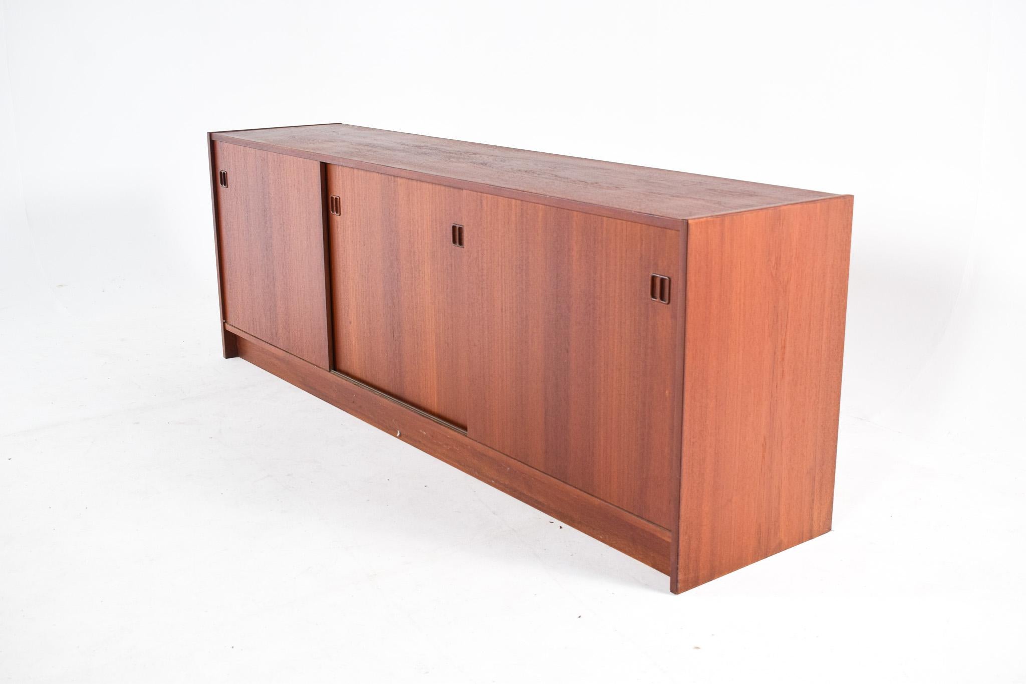 This image shows a quintessential mid-century teak sideboard that exudes the simplicity, warmth, and functional design characteristic of the period. The sideboard's long, low profile is typical of mid-century modern furniture, suggesting a subtle