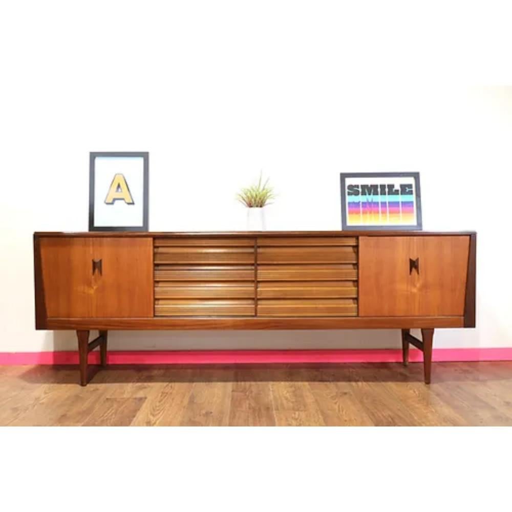 This Mid Century Modern Teak Vintage Sideboard Credenza by Elliots of Newbury is a stunning example of 1960s British furniture design. The teak wood has a beautiful grain that is sure to catch the eye of anyone who enters the room. The elegant