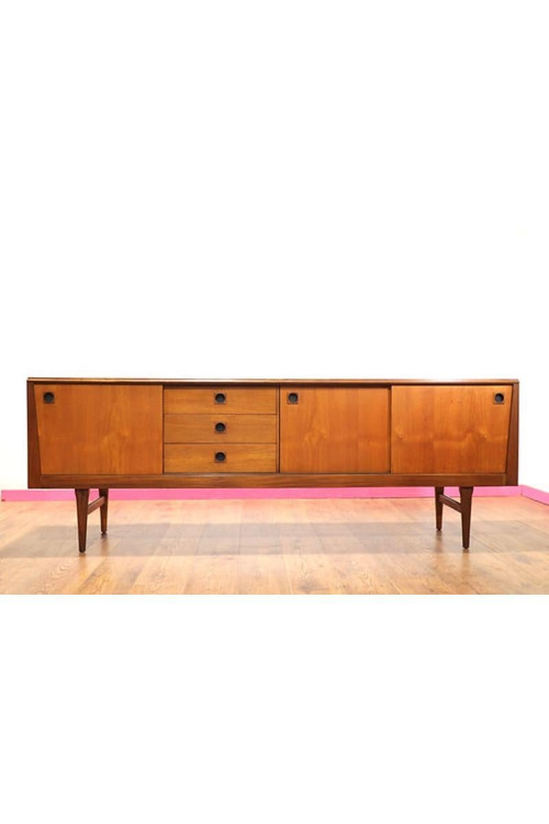 This Mid Century Modern Teak Vintage Sideboard Credenza by Elliots of Newbury is a stunning example of 1960s British furniture design. The teak wood has a beautiful grain that is sure to catch the eye of anyone who enters the room. The elegant