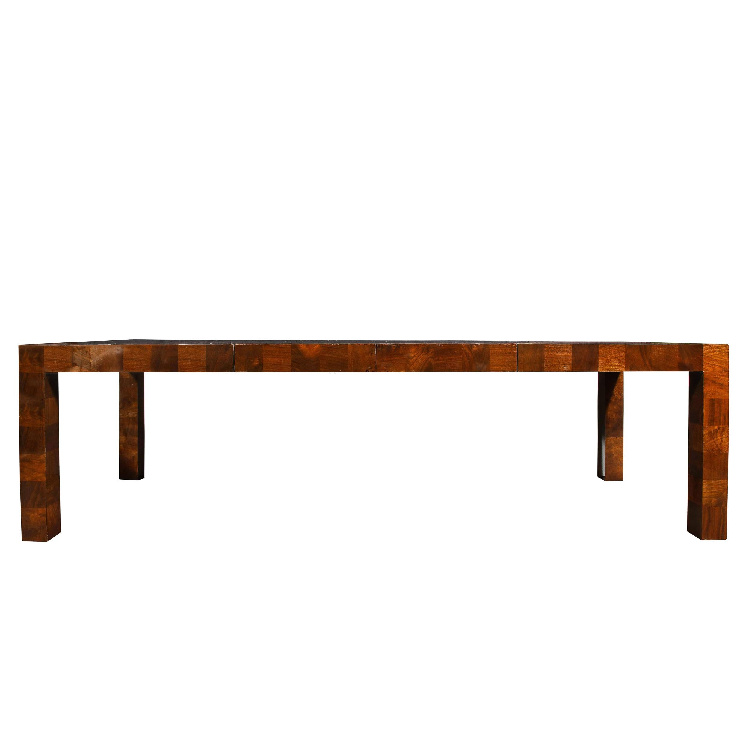 This gorgeous Mid Century Modern parsons style dining table was realized in the United States circa 1970. It features a rectilinear body features volumetric rectangular legs and a rectangular top in a parquetry mosaic of square tessellated burled
