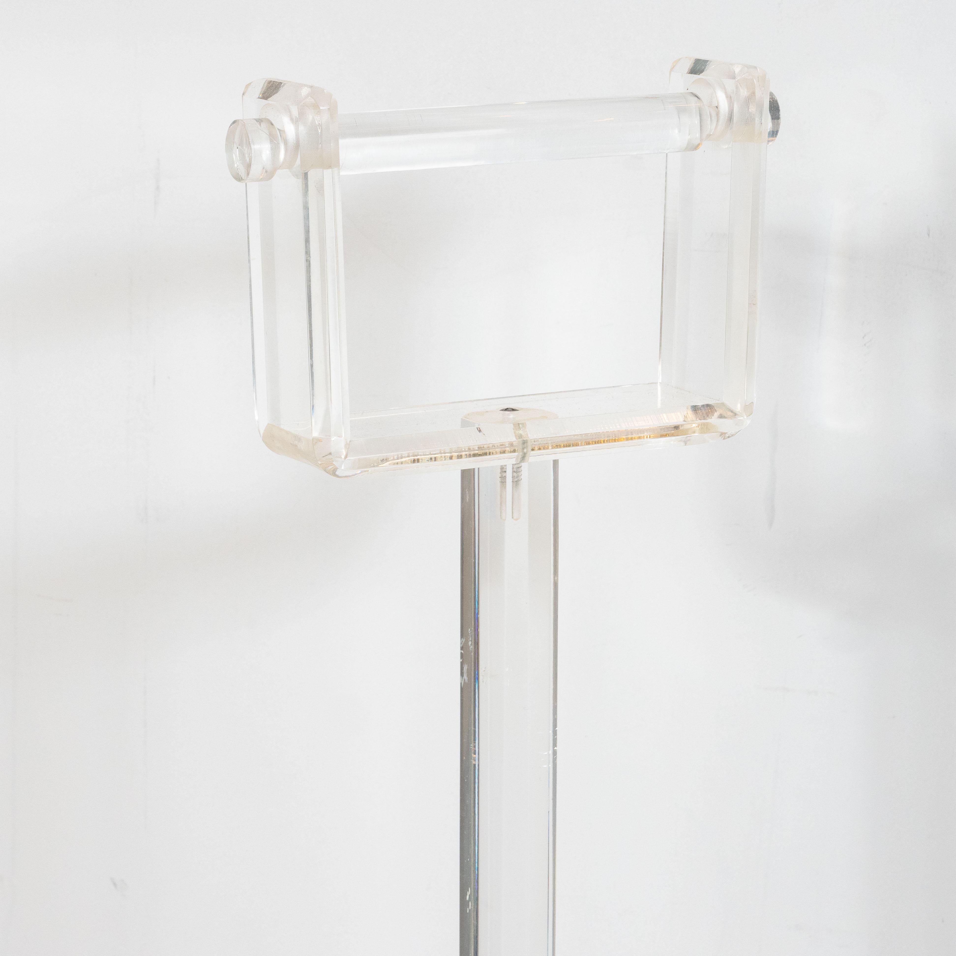 This sophisticated Minimalist toilet paper holder was realized in the United States circa 1970. It features a beveled base; an octagonal faceted rod that connects to a U-form embellishment at top speared through the center with a cylindrical