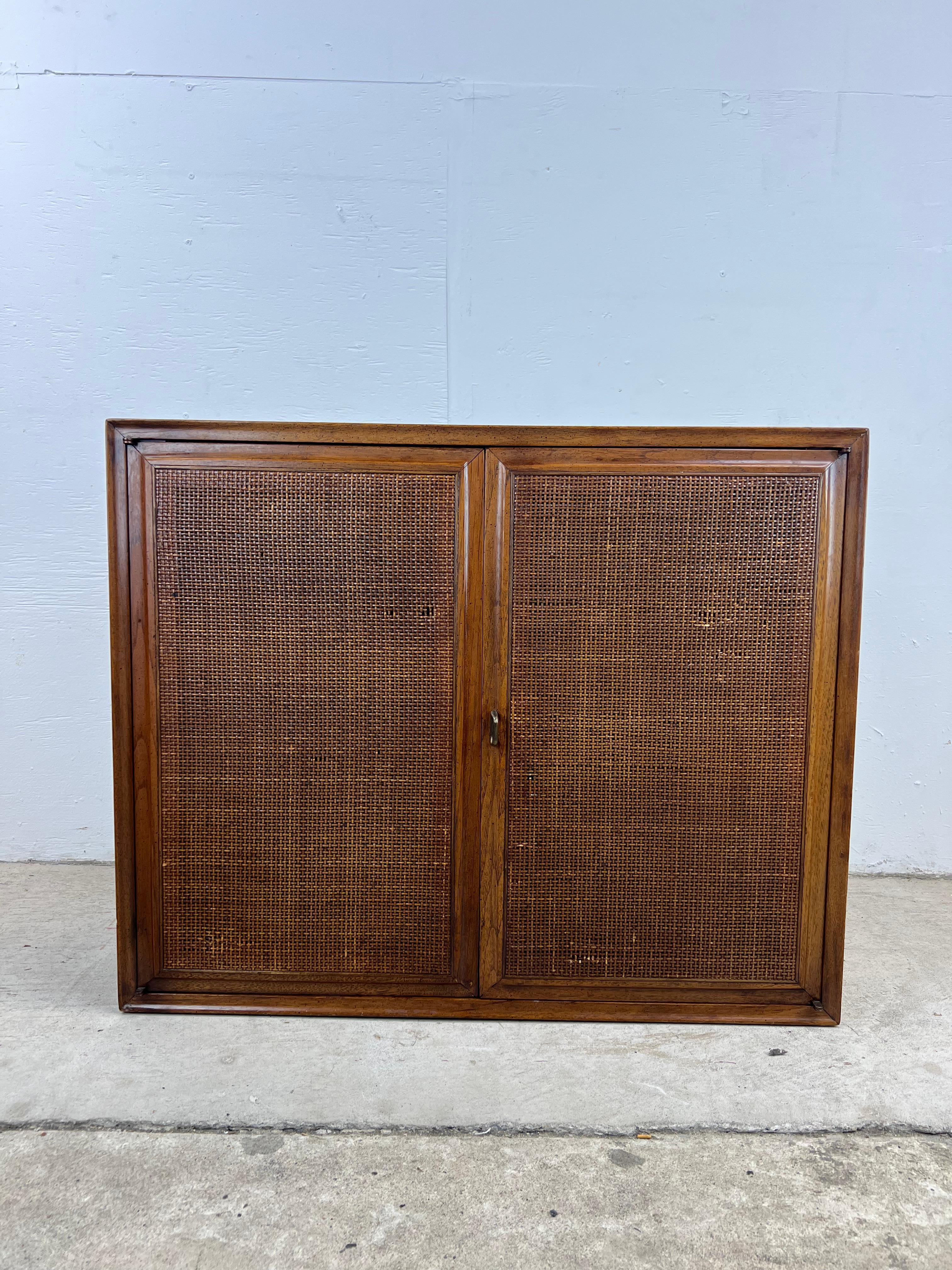 This mid century modern two door cabinet features pressed wood construction, walnut veneer with original finish, two cabinet doors with caned front, interior adjustable shelf, and brass accented hardware.

This cabinet appears to be by Jack
