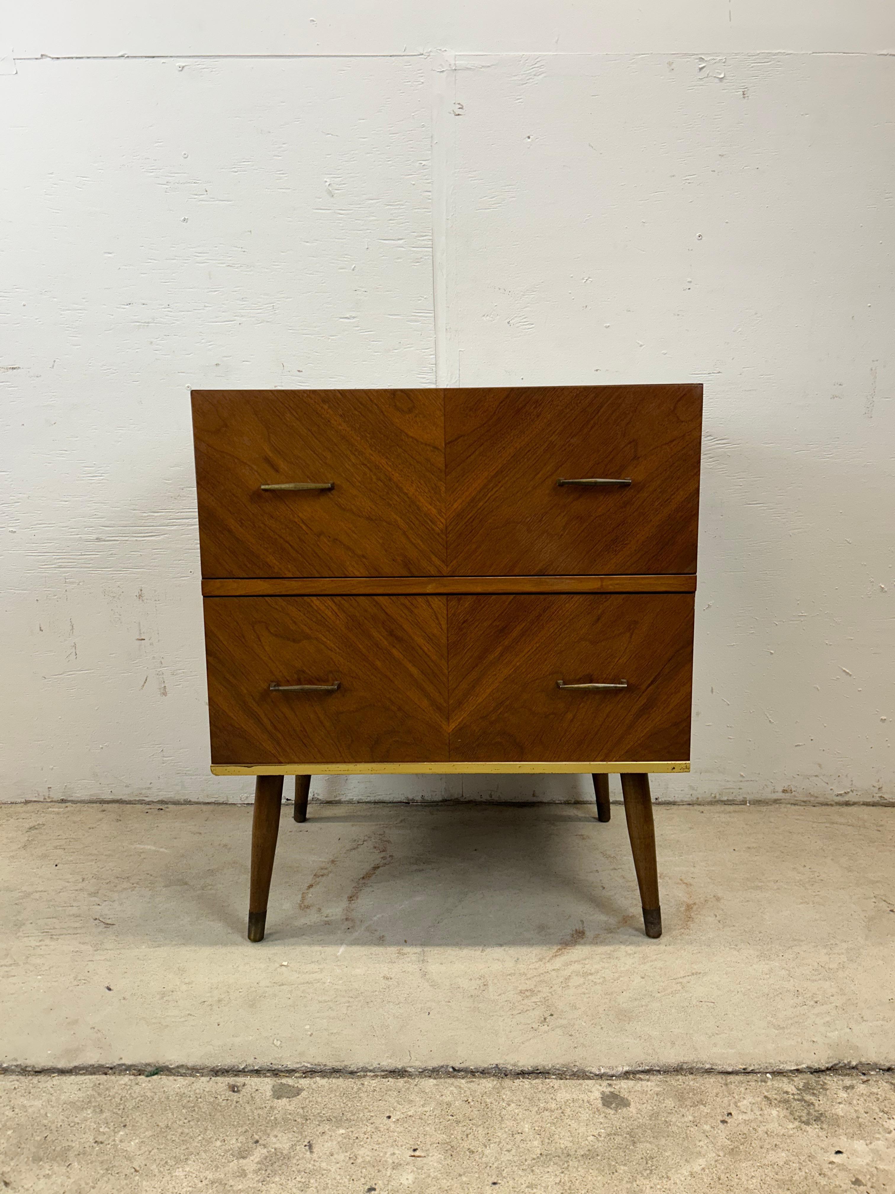 This mid century modern nightstand features hardwood construction, walnut veneer with original finish, two dovetail drawers with brass accented hardware, and tall tapered legs with brass capped feet.   Matching highboy dresser available