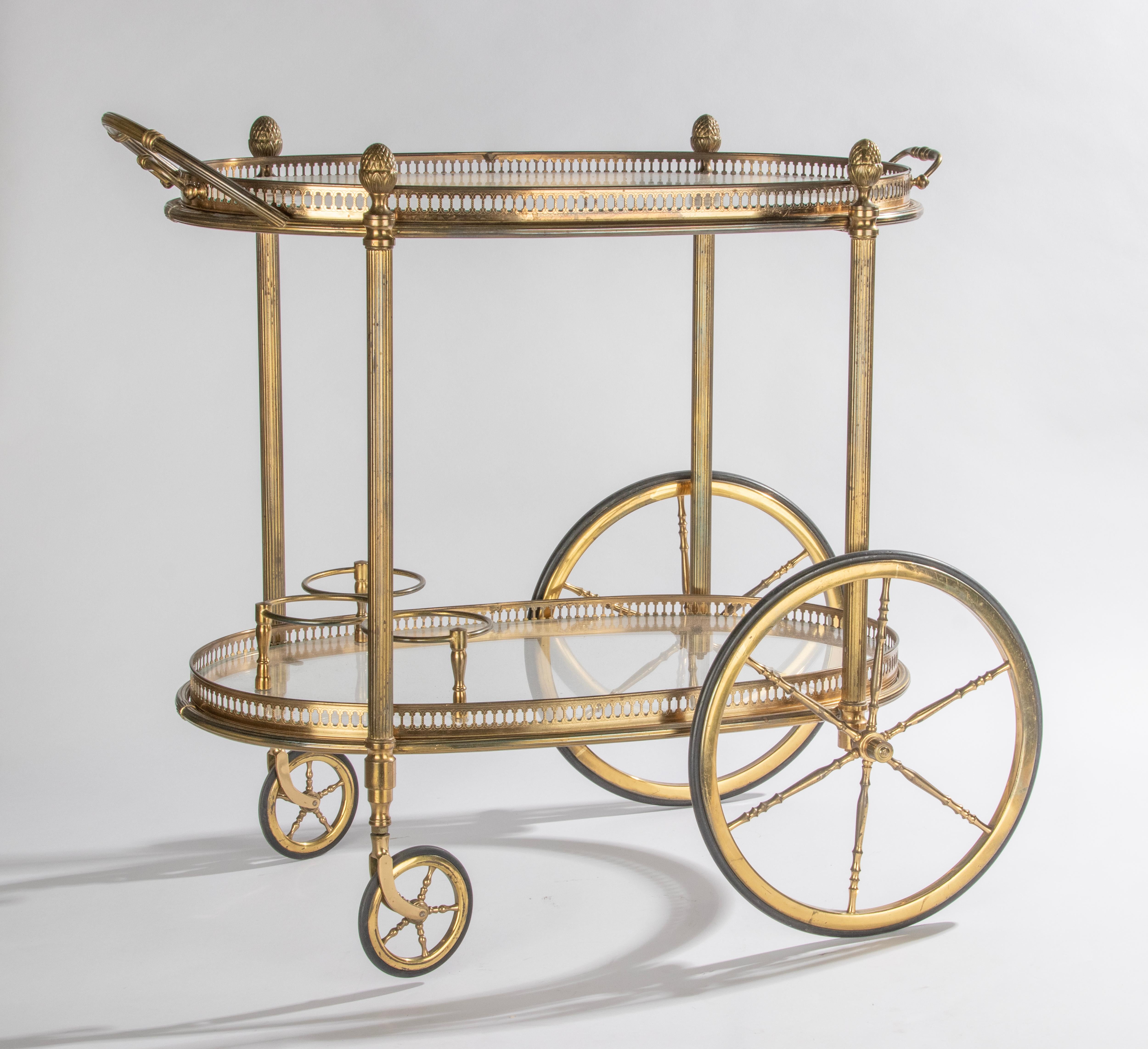Elegant chariot de bar à deux niveaux avec verre, attribué à la Maison Baguès, Paris. Le chariot est en métal et verre de couleur laiton, avec de beaux détails raffinés, comme les galeries en laiton percées, les poignées et les pommes de pin