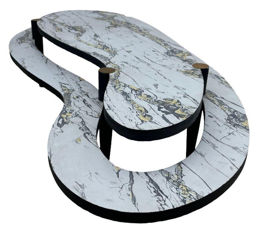 This biomorphic, Mid-Century Modern coffee table features two-tiers embellished with marbleized white Formica surfaces. The table rests on black-painted legs capped with brass foot details.