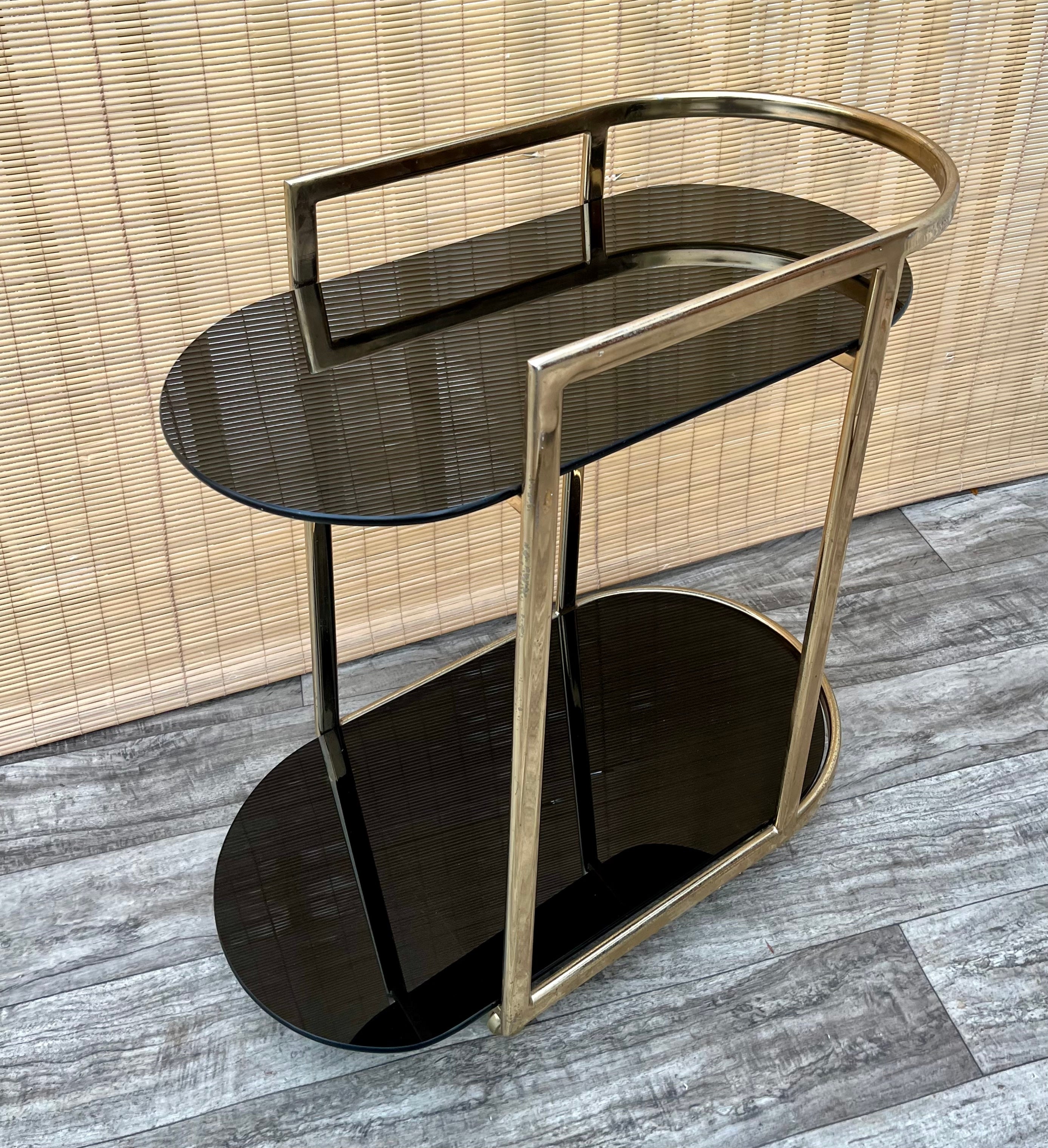 Vintage Mid Century Modern Two Tier Brass & Glass Dry Bar/ Serving Cart. Circa 1970s
Features a Hollywood regency inspired design with a brass plated frame, two tiers with rounded almost black-finish mirror glass shelves and casters for easy