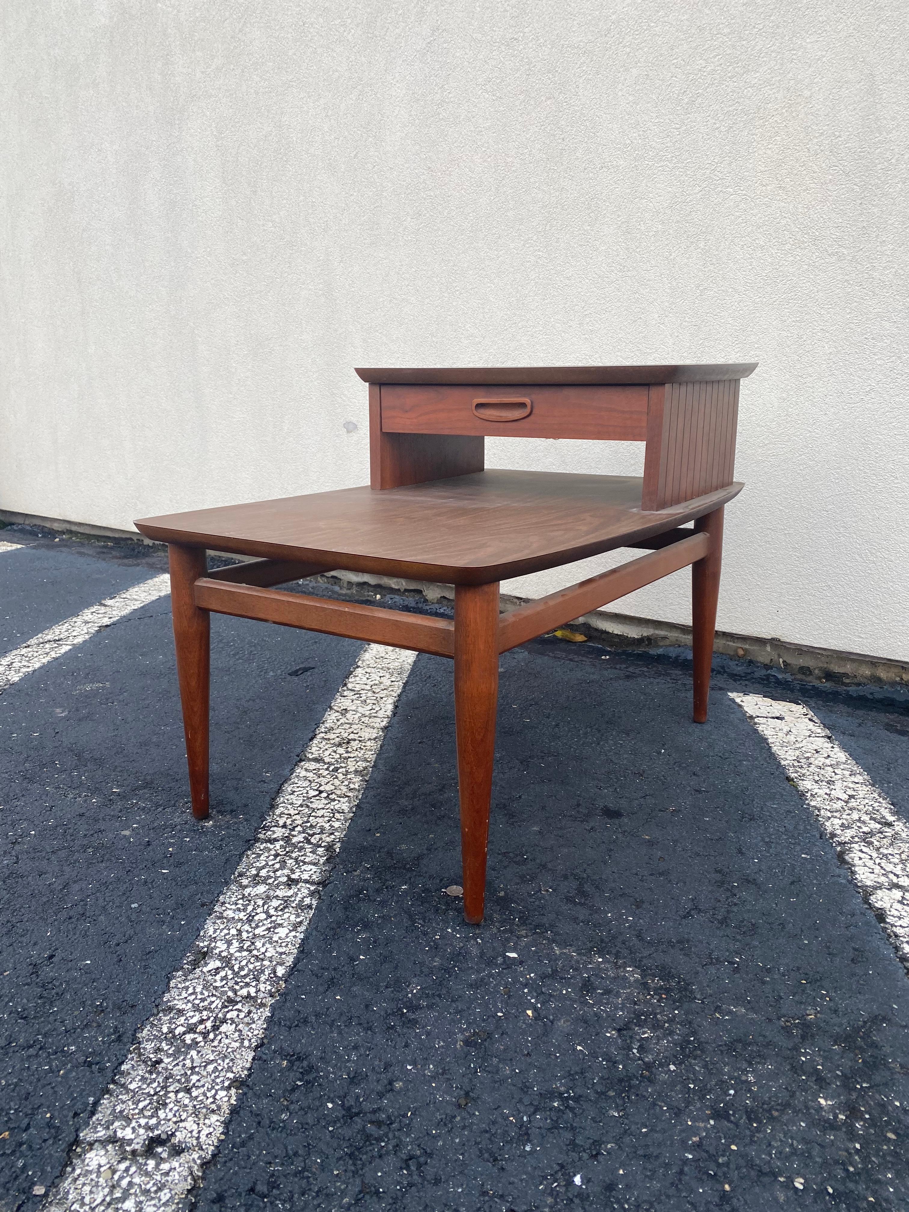 Mid-century two-tier lamp table by Lane. Walnut table with laminate top and drawer with recessed pull. Beautiful wood tone and tapered legs with vertical reed detail at side.