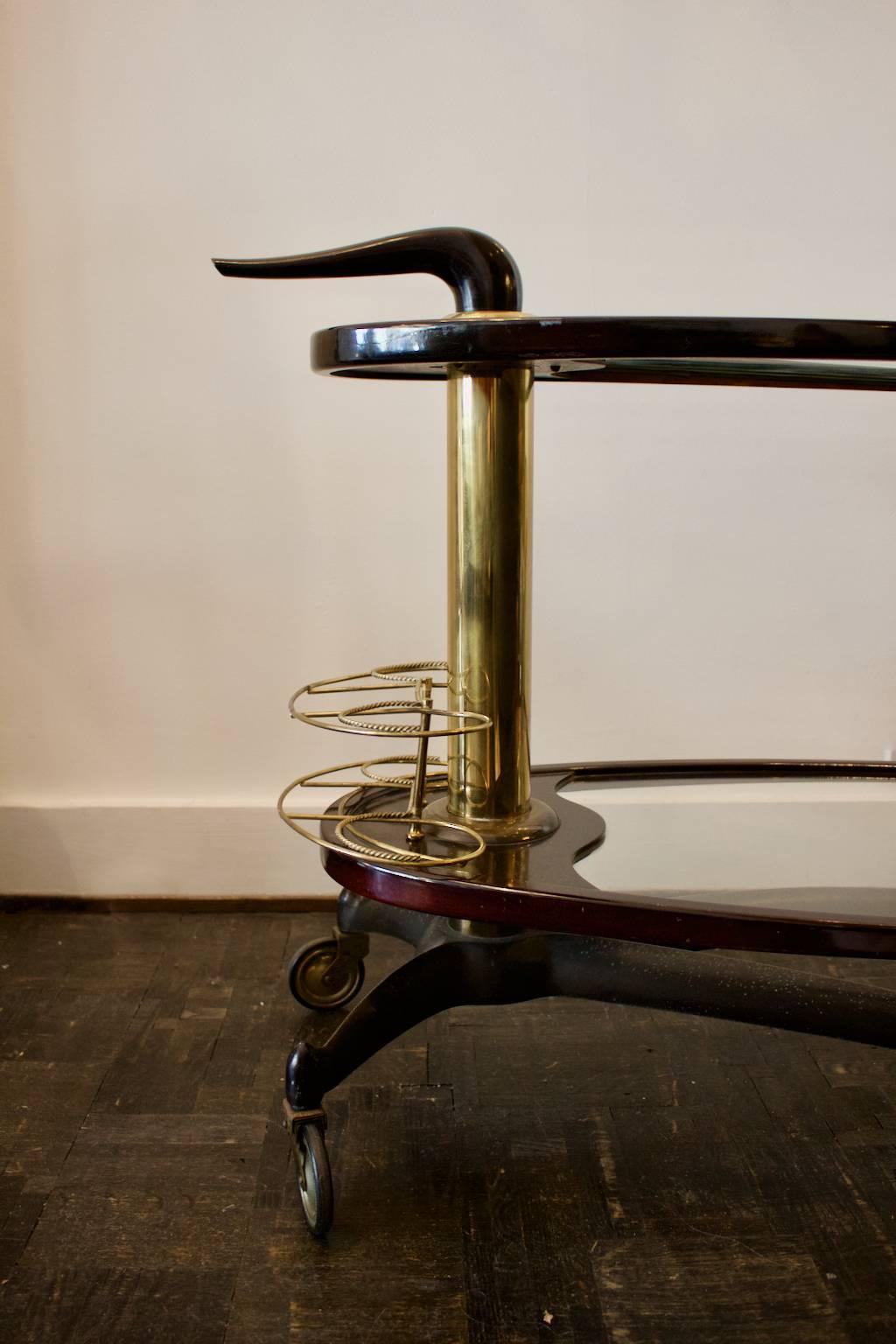 An elegant wooden bar trolley with two cantilevered tiers and brass details, Italy, 1950s.

The trolley is constructed around a steel X-shaped lower frame with four wheels and heavy brass column, and twin shelves framed in wood (probably