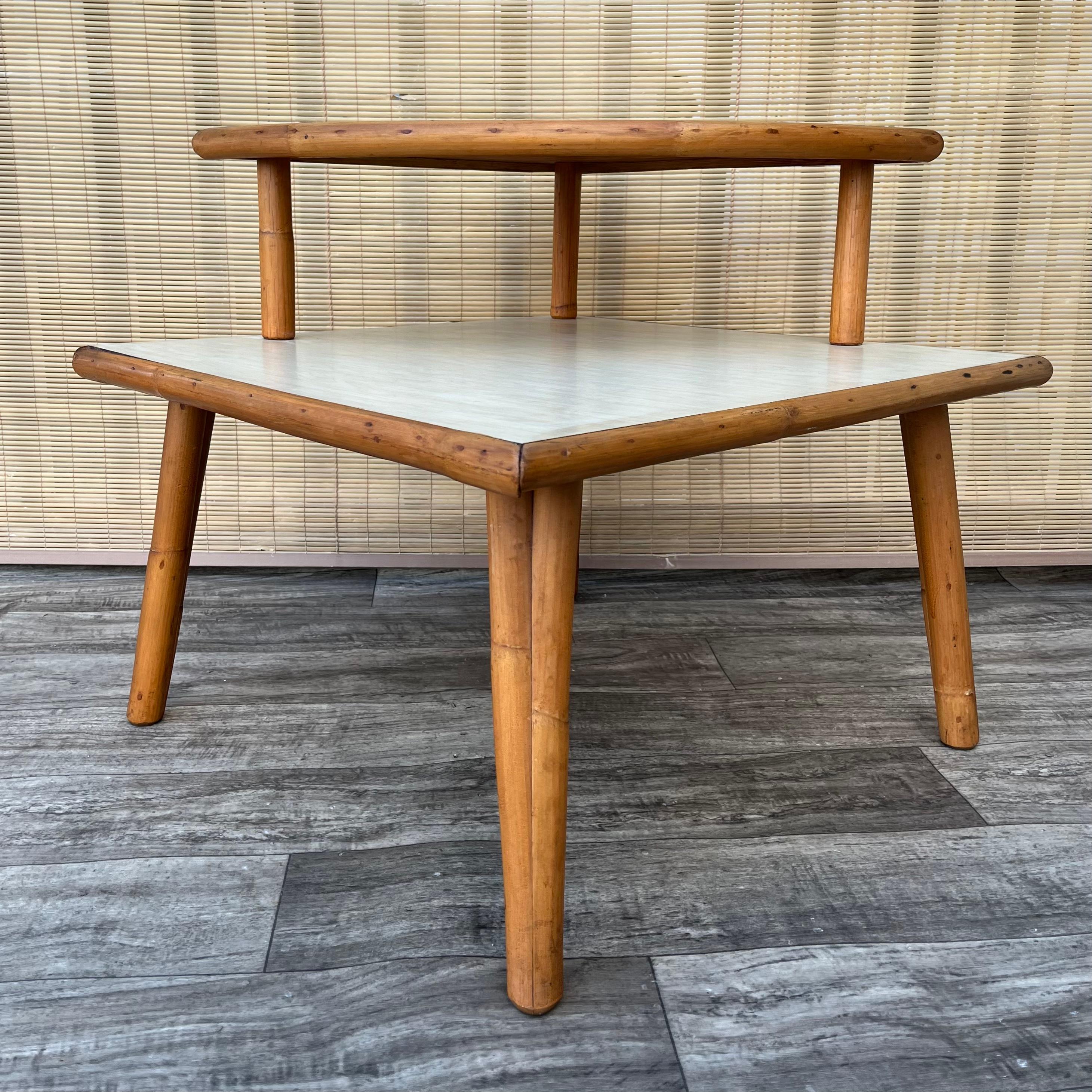 Vintage Mid Century Modern Two Tiered Rattan Corner Table in the Paul Frankl's Style. C 1950s
Features a quintessential Mid Century Modern Design, two tiers with cream color laminated tops, and a sleek rattan frame.
In good original condition, with