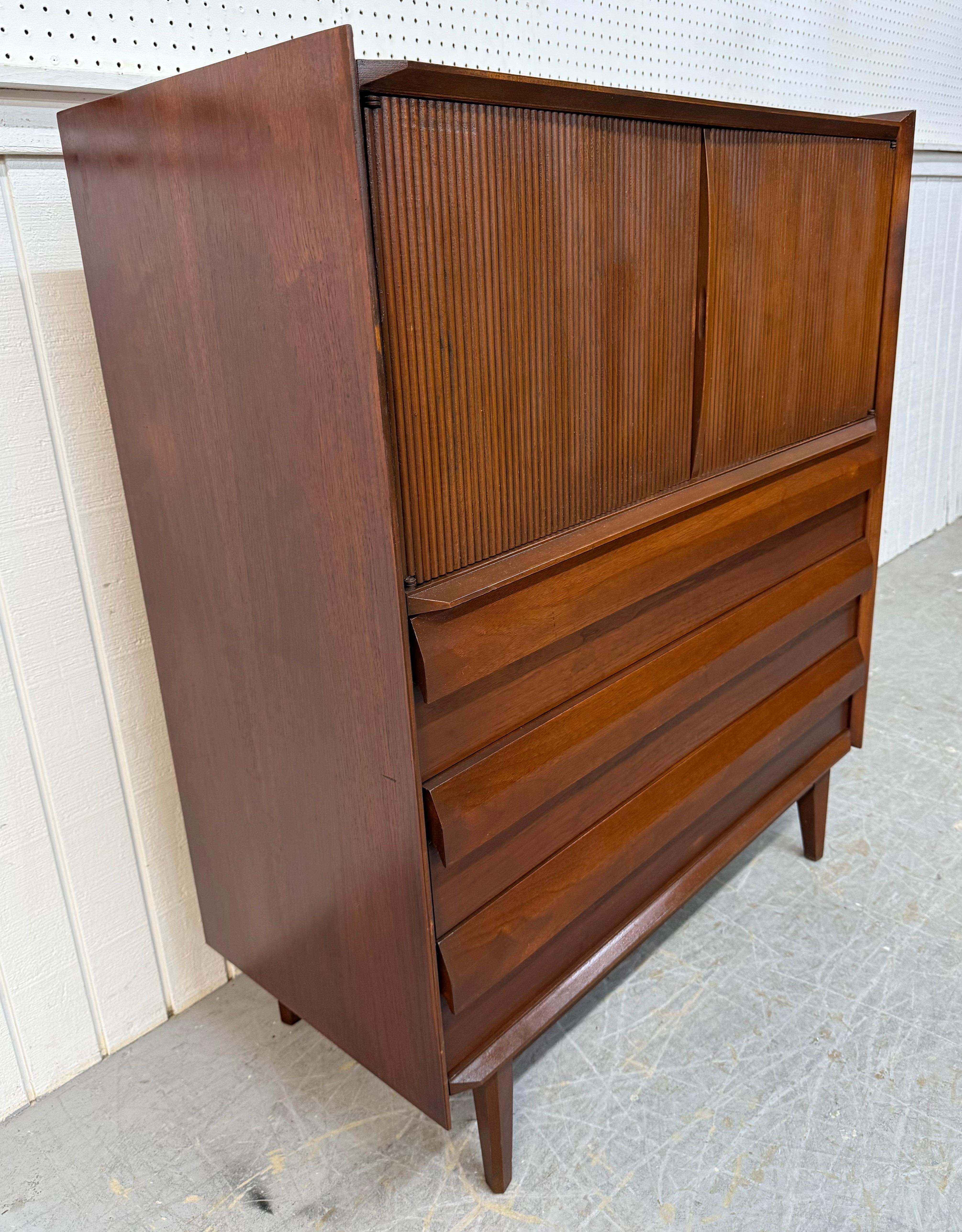 Cette annonce concerne un meuble haut en noyer de la collection United 1ST Edition, de style moderne du milieu du siècle dernier. Elle se caractérise par un design rectiligne, deux portes avant cannelées qui s'ouvrent sur deux tiroirs cachés, trois