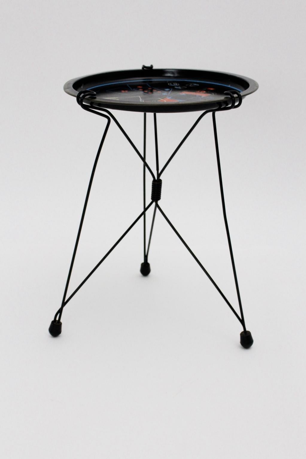 This Mid-Century Modern vintage side table shows a round black lacquered sheet metal top with multicolored motifs. The black lacquered wire steel base is foldable and features rubber feet.
The vintage condition is good with signs of age and