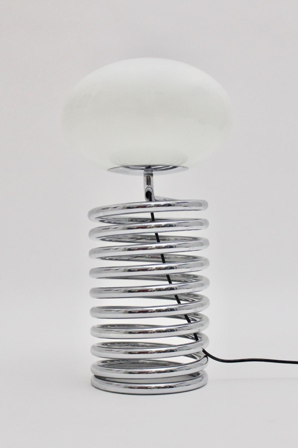 This spiral or loop table lamp was designed by Ingo Maurer 1968 Germany for Design M. Germany.
The table lamp features a tabular chromed metal spring as a base and an opaline glass shade. Also the table lamp shows one E 27 socket and an on/off