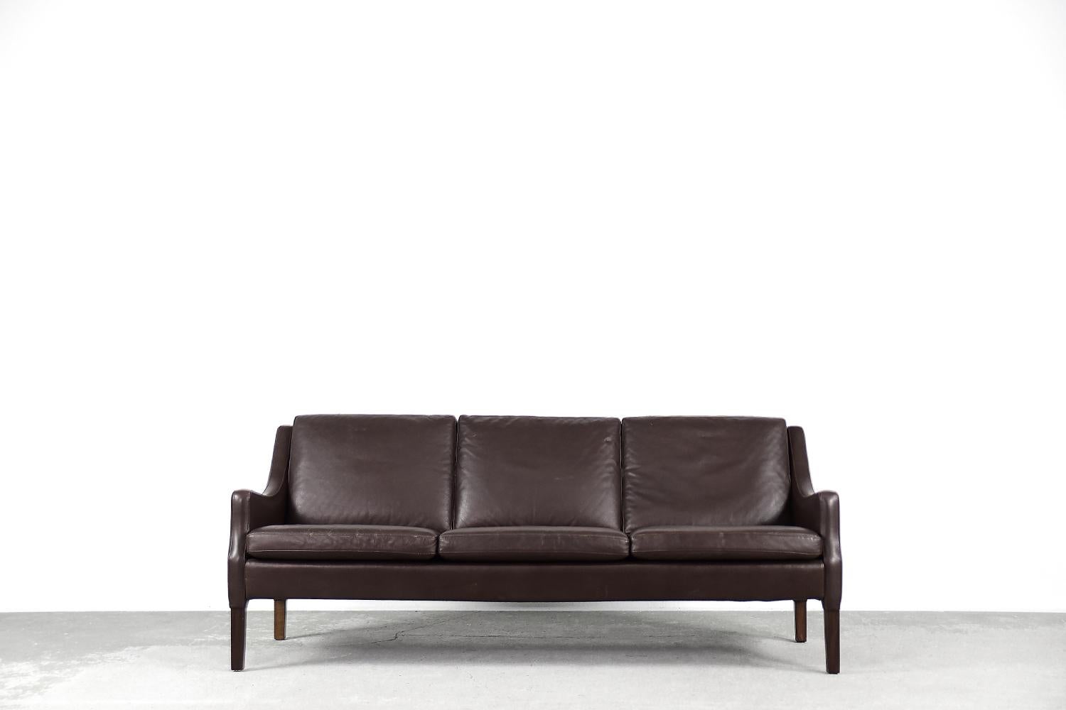 This three-seater sofa was made in Denmark during the 1960s. The sofa is completely upholstered in natural leather in a deep dark chocolate color. Comfortable, loosely placed cushions ensure comfort and convenience during use. The legs are made of