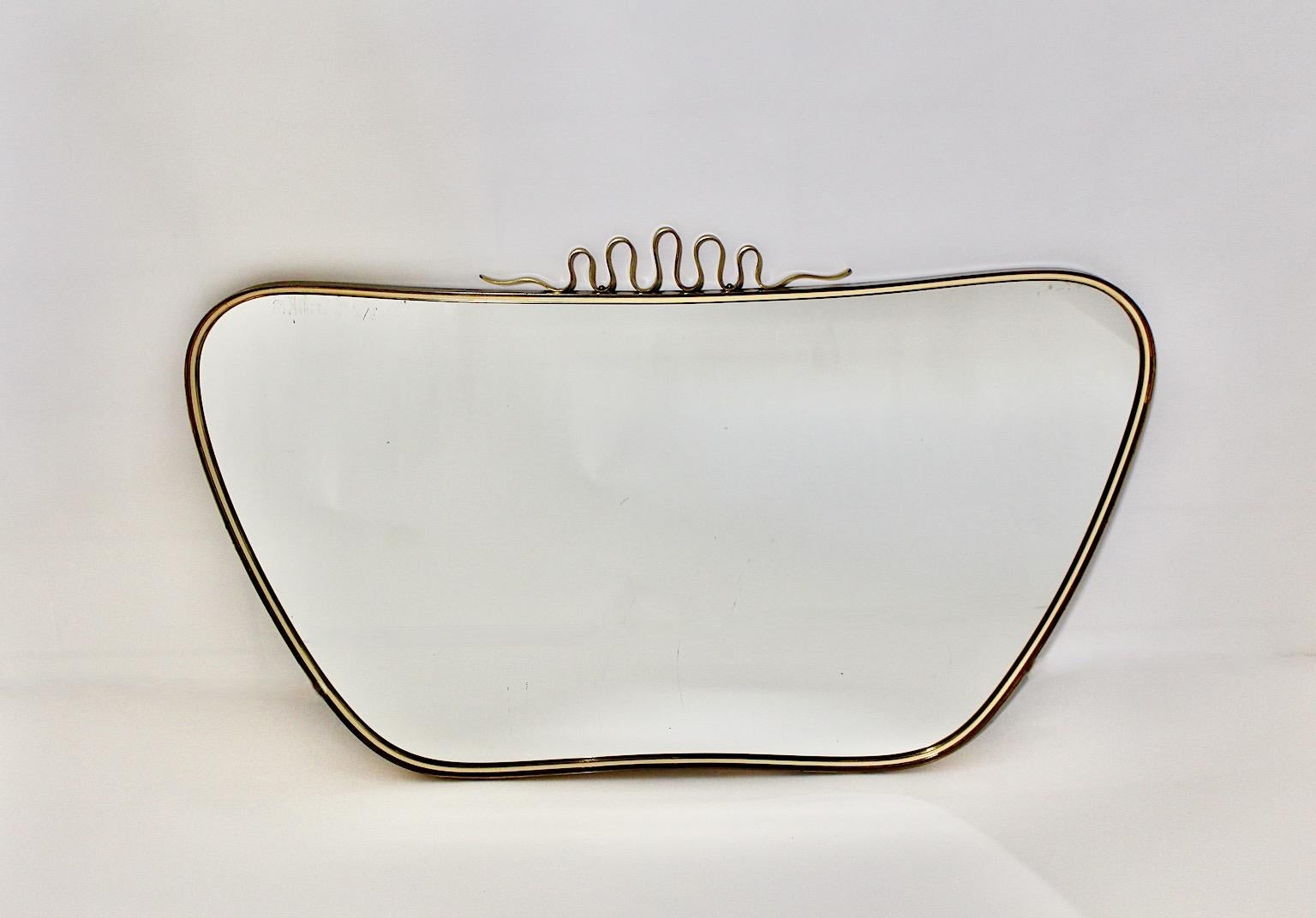 Mid-Century Modern vintage wall mirror in longish oval form with fabulous brass loops as decor at the top.
While the brass decor at the top shows amazing brass patina the mirror frame features a white framed line, which highlights the elegant and