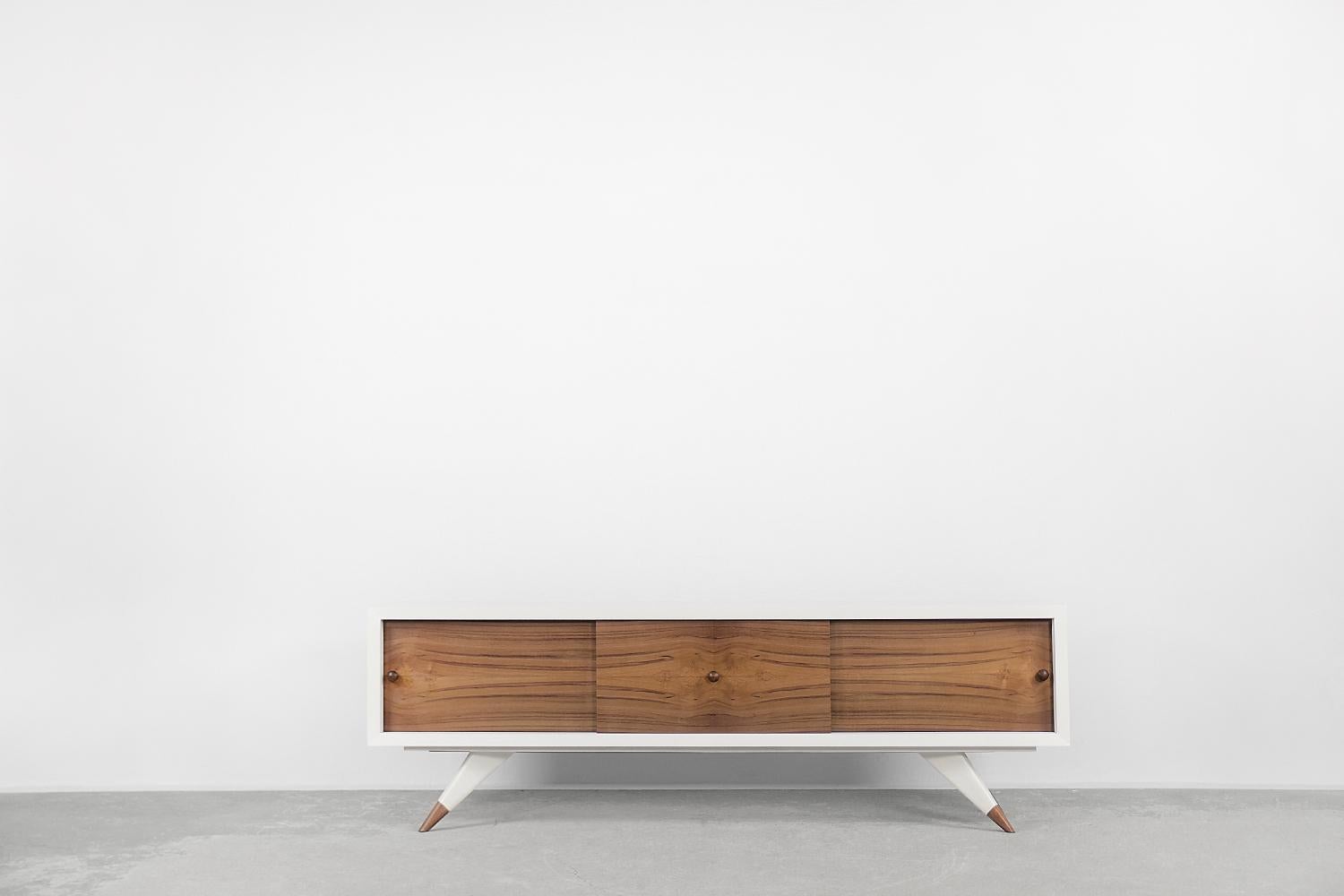 This modernist sideboard was manufactured in Sweden in the 1970s. It was finished with white lacquered teak wood. The front consists of three sliding doors in a natural shade of teak wood. Inside there are shelves and a spacious, empty space. The