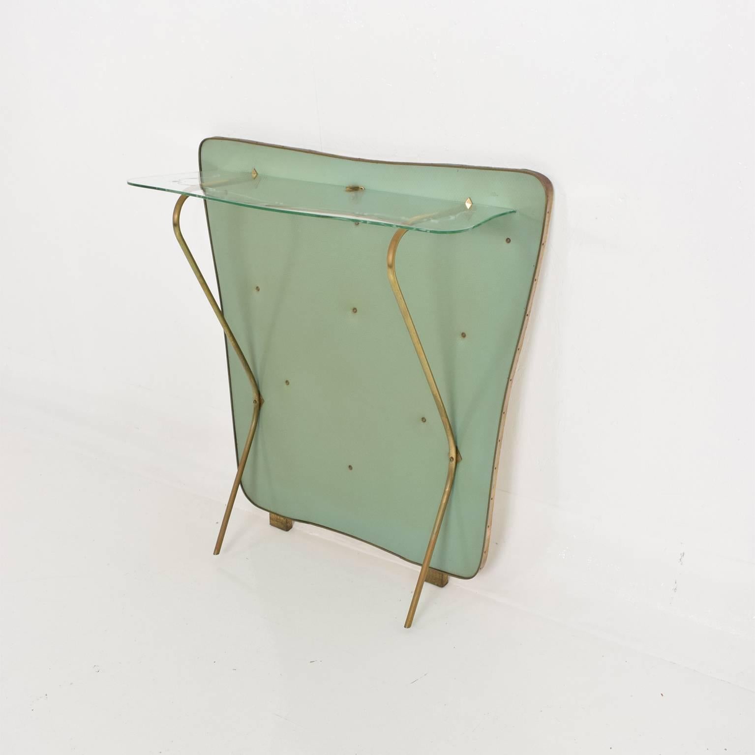 For your consideration a Mid-Century Modern wall console. Made in Italy, circa late 1950s.
Wood board with original green vinyl cover. Brass frame, legs, and brackets support the sculptural original glass.

Dimensions: 32 1/2
