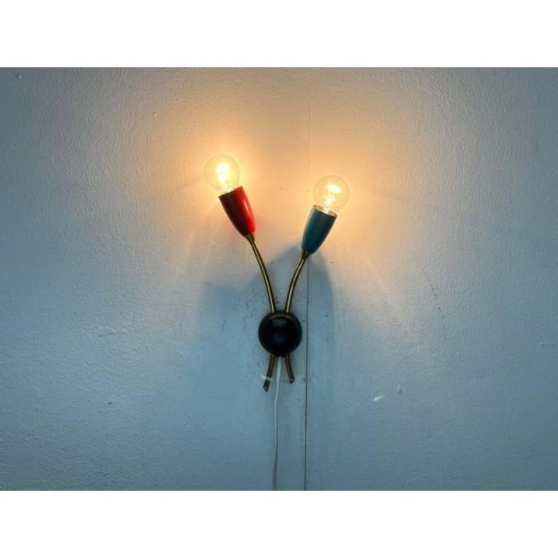 Mid-century wall light with a distinctive color scheme.

At the end of two elegantly curved brass rods sit the sockets with cup-shaped shades. The shades are colored red and blue-green.

The wall base forms a black hemisphere. The two brass shafts