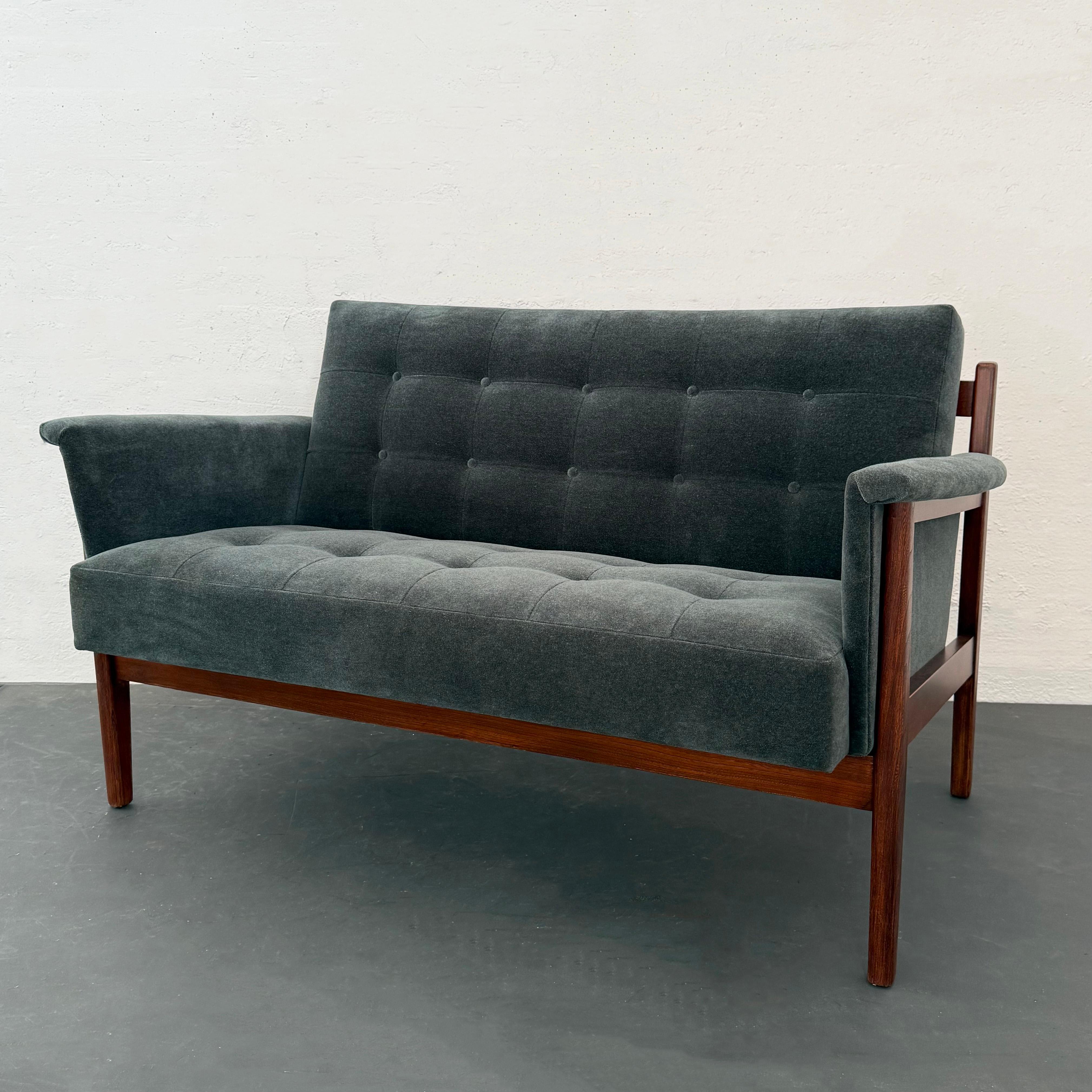 This classic Mid-Century Modern loveseat settee by the Paoli Chair Company in Orleans, Indiana circa 1965 features a solid walnut frame with an exposed horizontal back slat. It is newly upholstered in a soft and cozy steel blue chenille with tufted