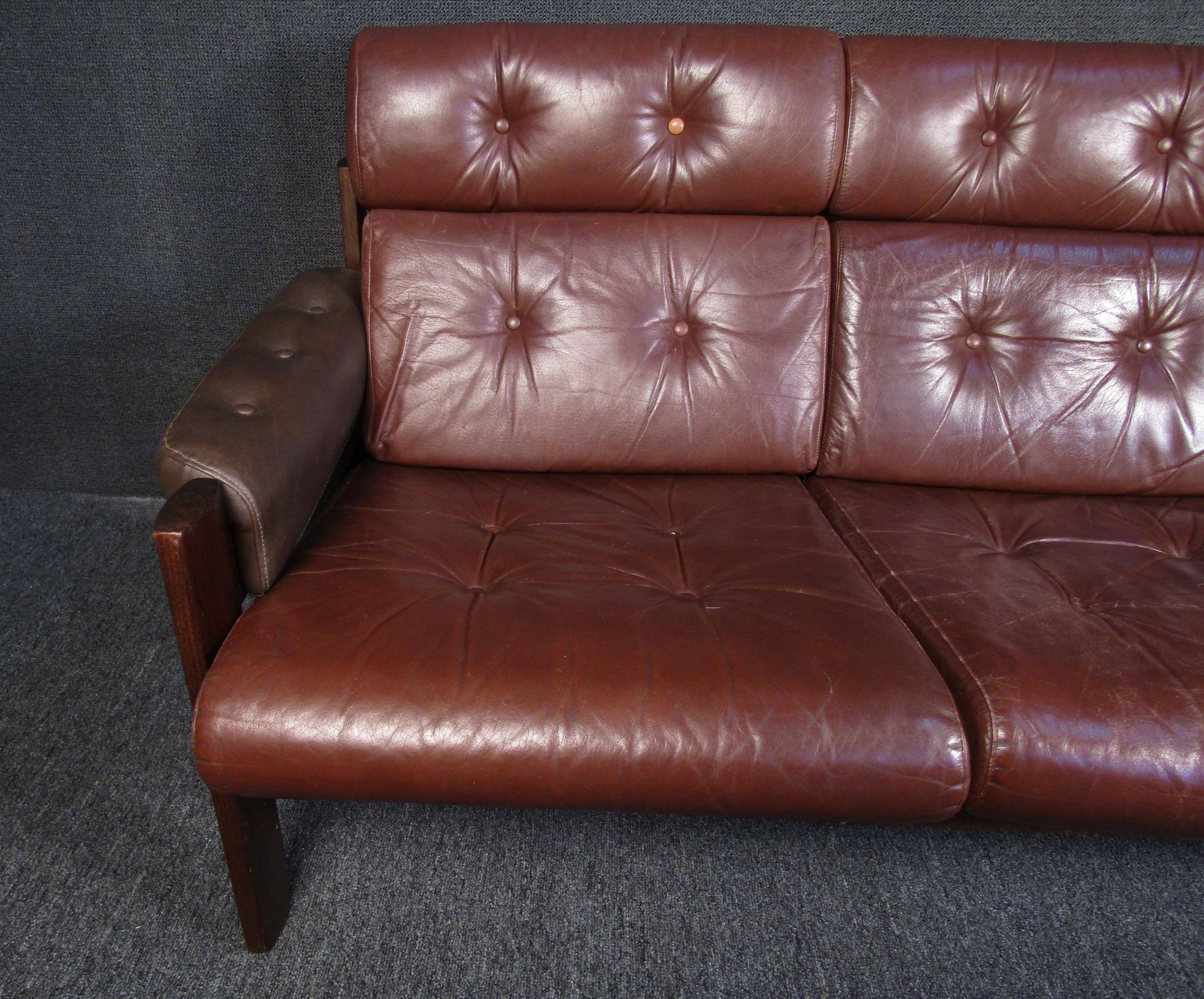 A beautiful midcentury couch that is sure to wow with its quality construction of leather and walnut wood. Burgundy leather covers most of the couch's surface while a darker shade of brown leather covers the couch's arms. The frames and legs