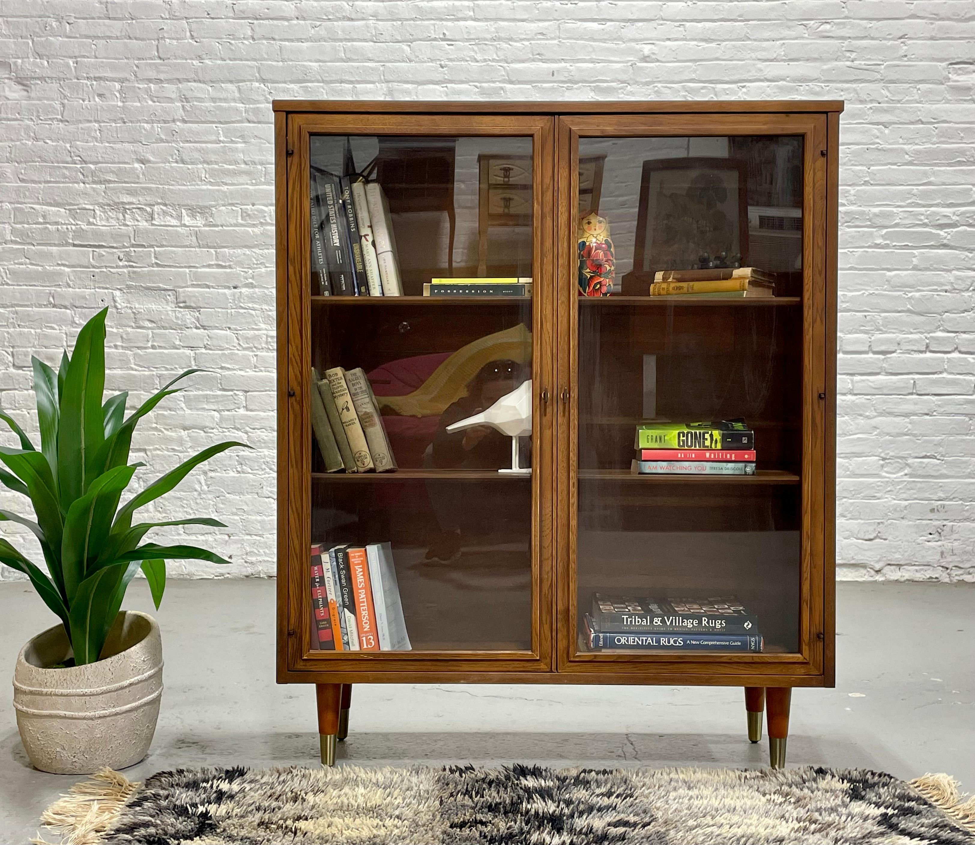 Mid Century Modern Walnut Bookcase / China Cabinet, c. 1960’s.   Three rows of generous shelving provides loads of space for all your books or to display your collection. Glass doors help keep the dust out and the brass capped tapered legs offer a