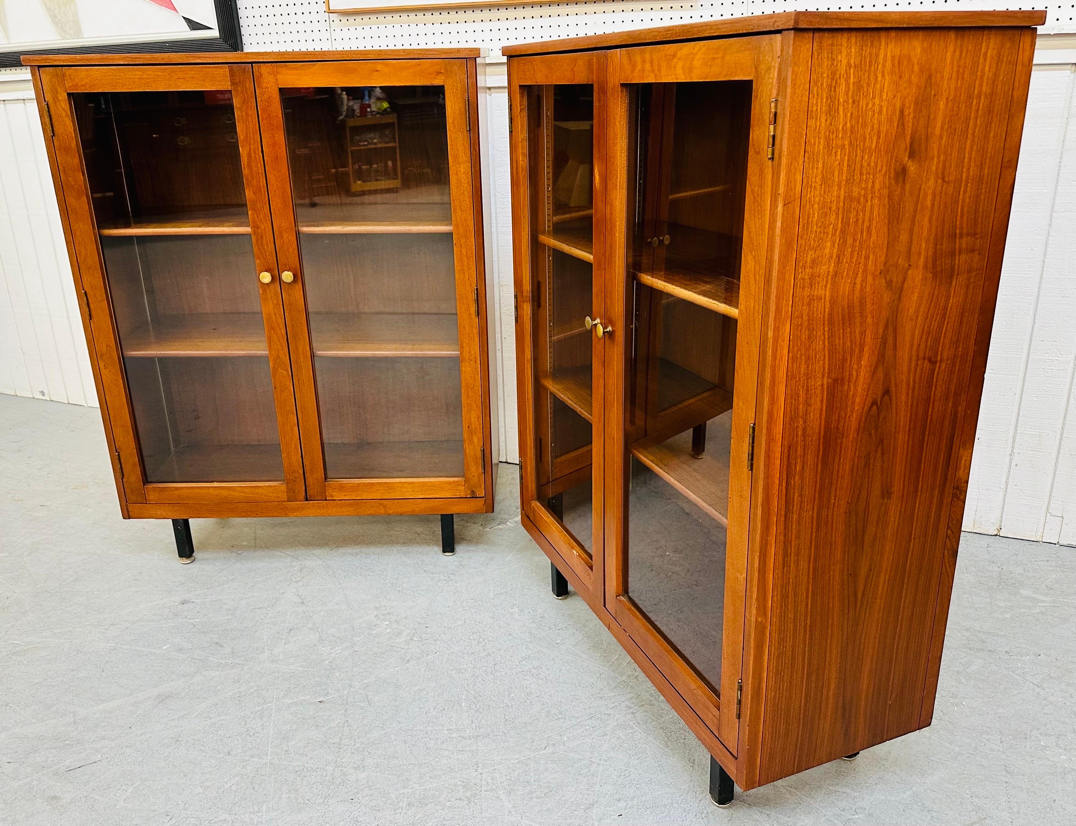 This listing is for a pair of Mid-Century Modern walnut bookcases. This matched pair features glass doors, two adjustable shelves, original pulls, black industrial legs, and a walnut finish.