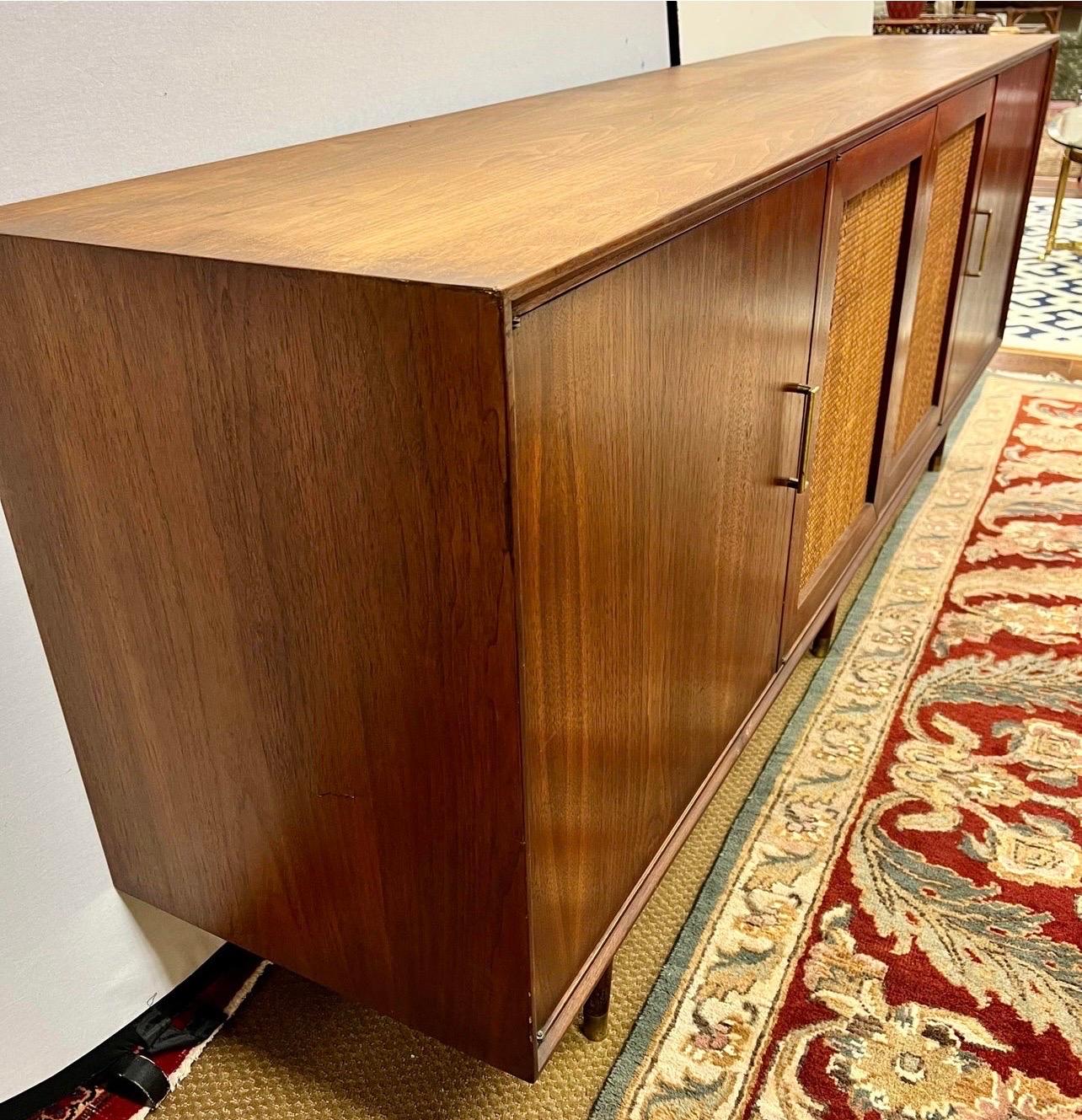 Handsome mid century credenza cabinet that has four doors and feature center doors that have caning. Side doors open to shelving and drawers and there is plenty of storage space. Decorative brass stretcher bar is a nice touch between the front legs.
