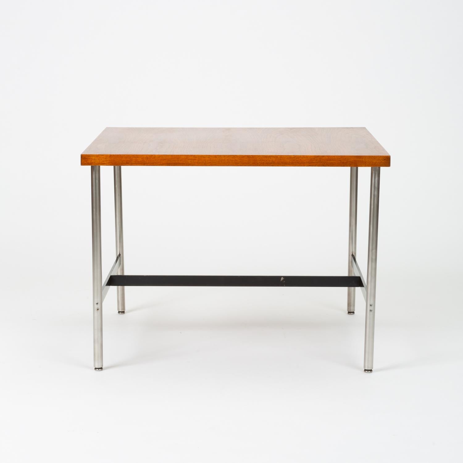 A modernist children’s table by Herman Miller with a square tabletop and metal frame. Four round legs of brushed steel with steel glides sit slightly inset from the corners of the work surface. An I-shaped metal support in black-enameled steel joins