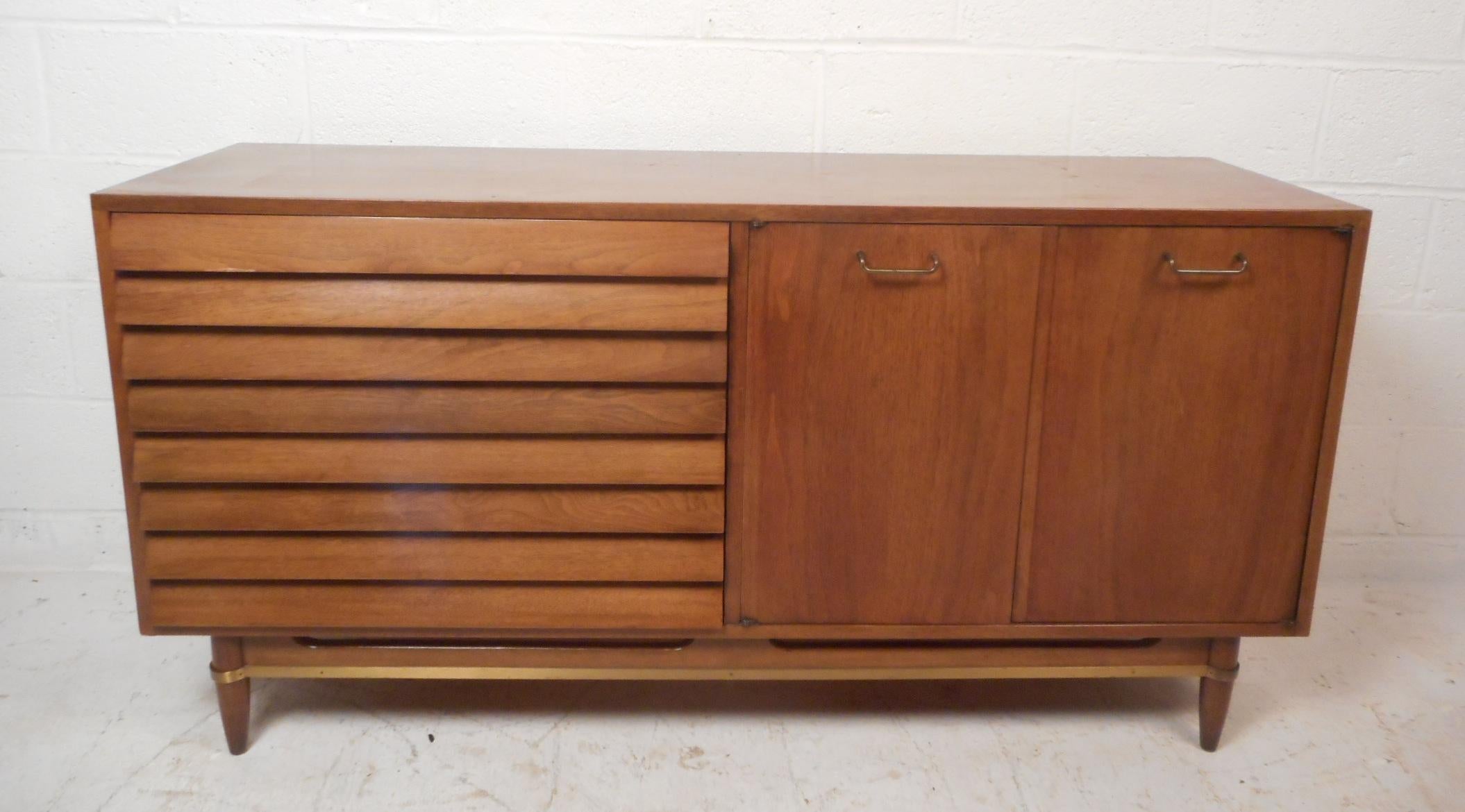This stunning vintage modern credenza features three large drawers with louvered fronts and three small drawers hidden by two cabinet doors. A fabulous case piece with a large hidden compartment, tapered legs, and a sculpted base with brass trim