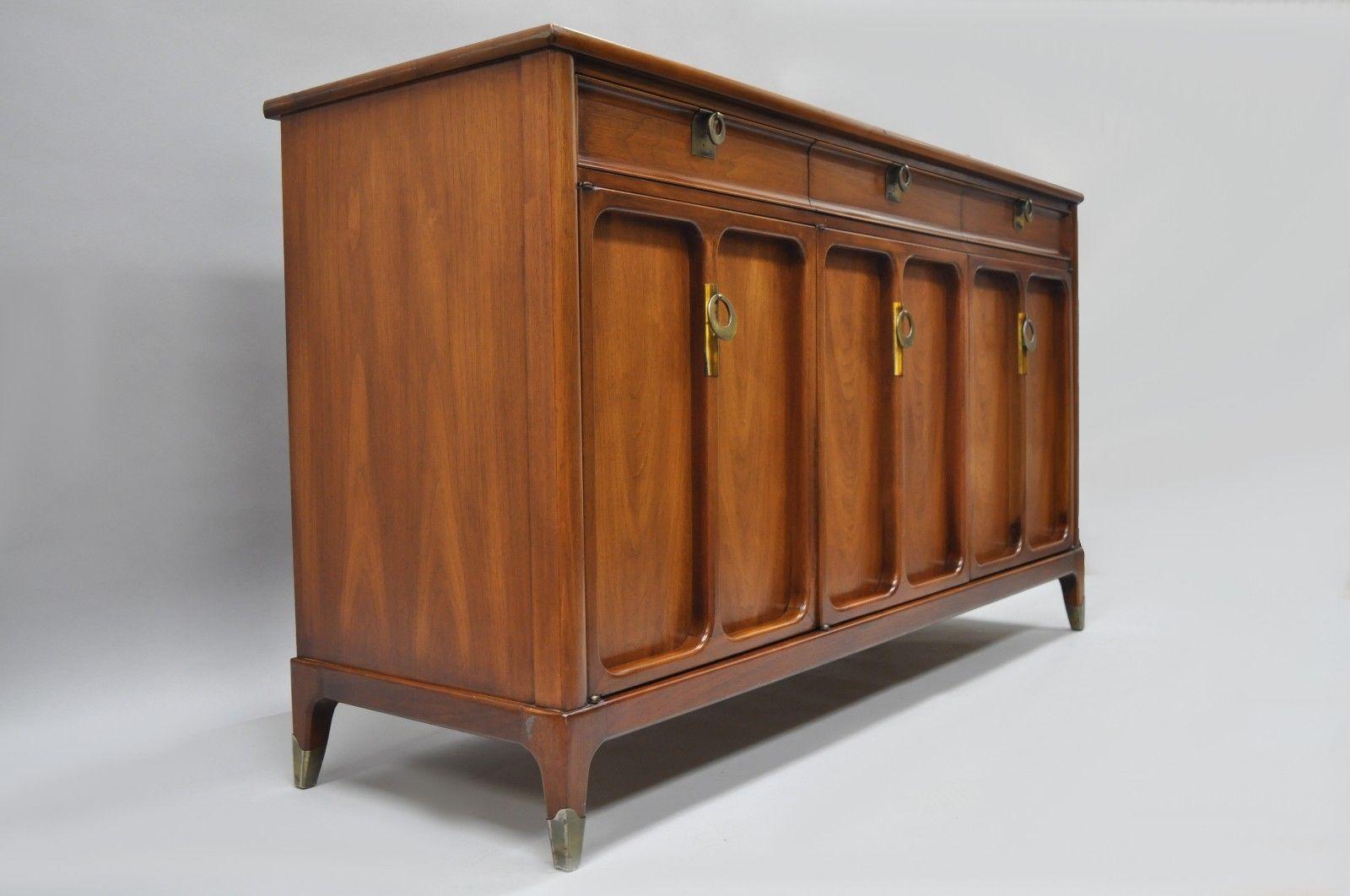James Mont style walnut sideboard by White Furniture Company. Item features beautiful woodgrain, three swing doors, three dovetailed drawers, two wooden shelves, tapered legs, brass capped feet, unique drop pulls, clean modernist lines, quality