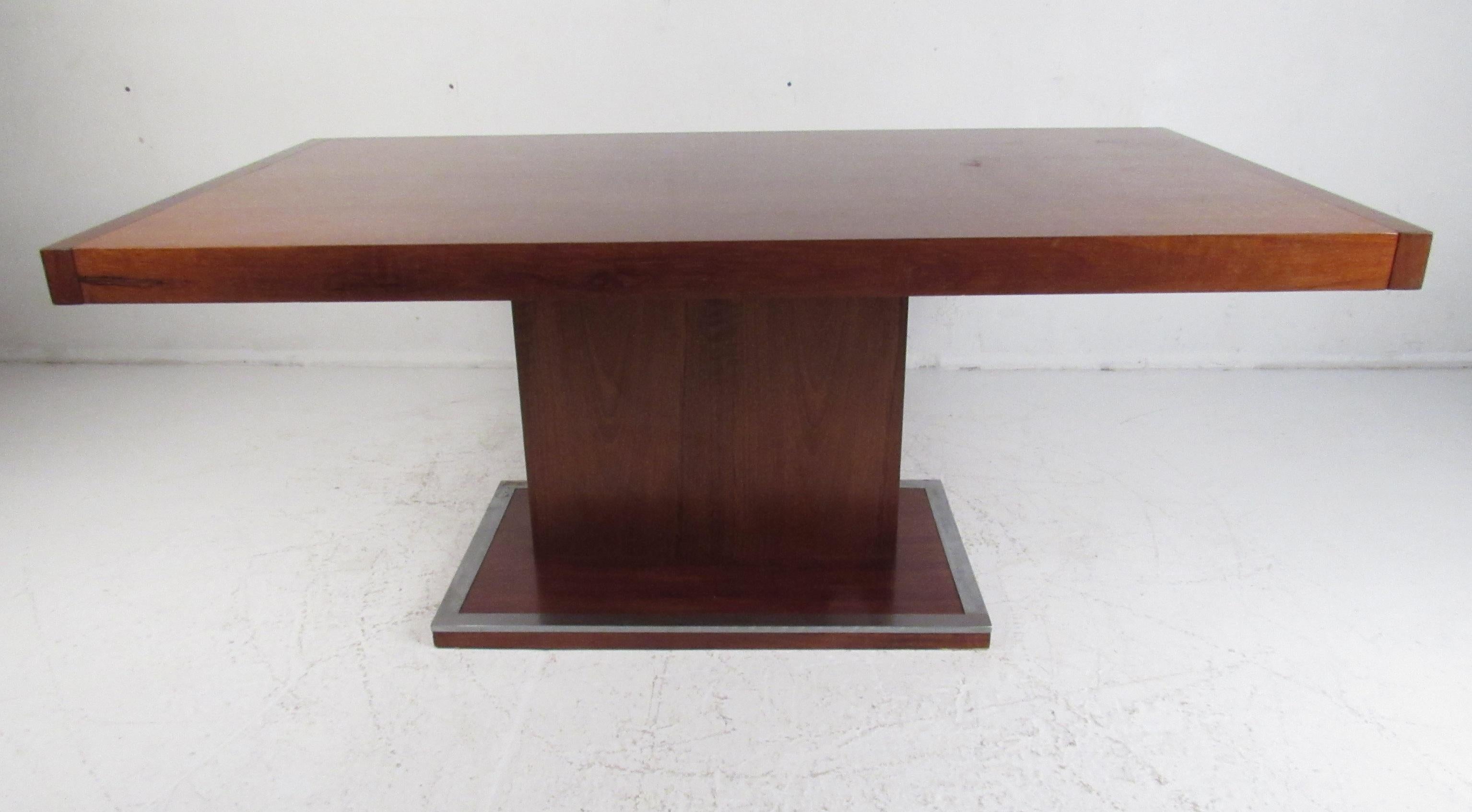 This stunning vintage modern dining table features a unique pedestal base with metal trim wrapped around the edges. The Art Deco style design, clean lines, and elegant walnut wood grain shows quality craftsmanship. This attractive mid-century dining