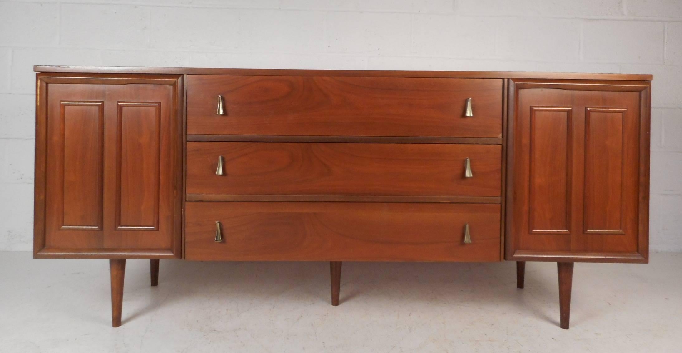 This stunning vintage modern credenza features six hidden drawers behind two cabinet doors and three large drawers in the centre. Unusual sculpted metal drawer pulls, tapered legs, and an elegant vintage walnut finish show quality craftsmanship. A