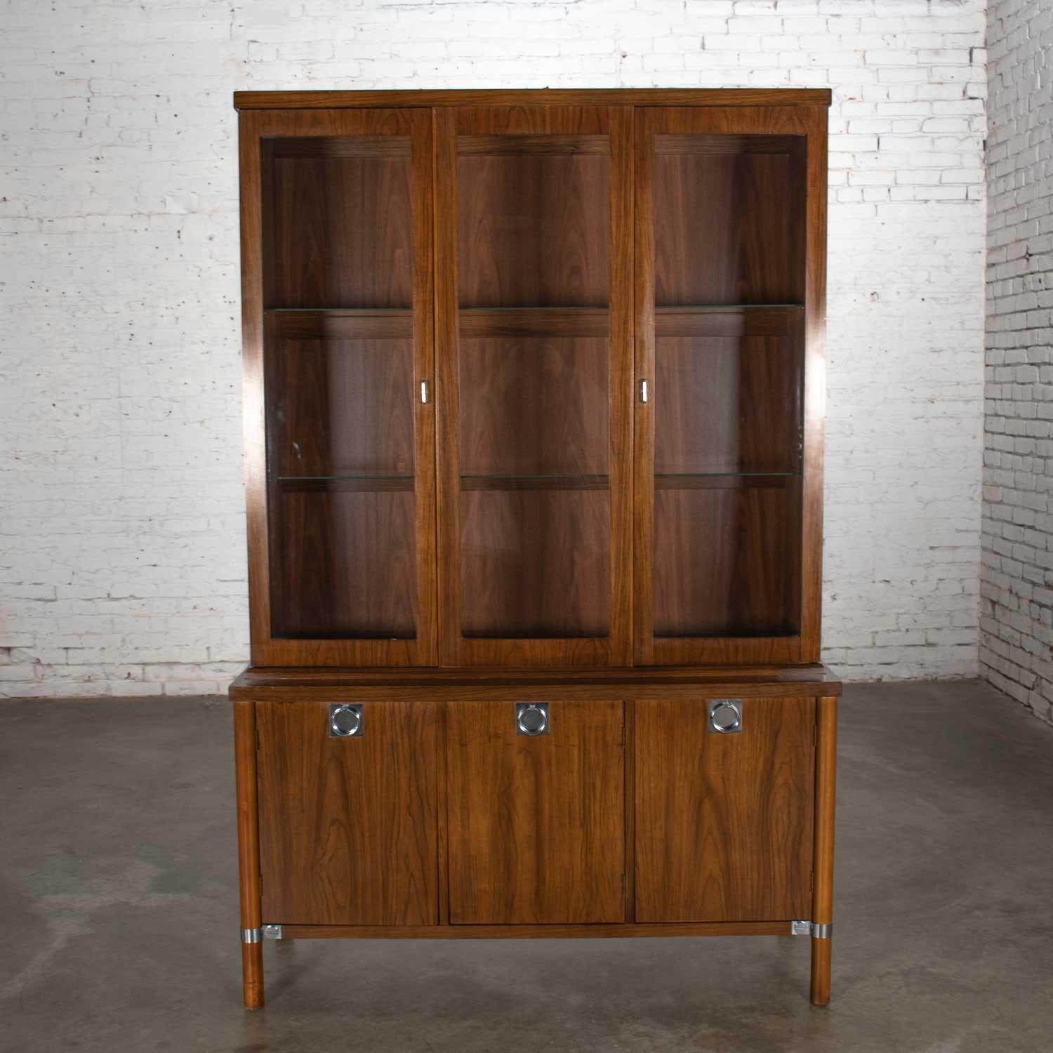 Gorgeous Mid-Century Modern walnut veneer china cabinet with chrome accents, glass shelves, and lights. Nice original vintage condition with wear as you would expect with age. One leg was chipped at the bottom and has been repaired and touched up.