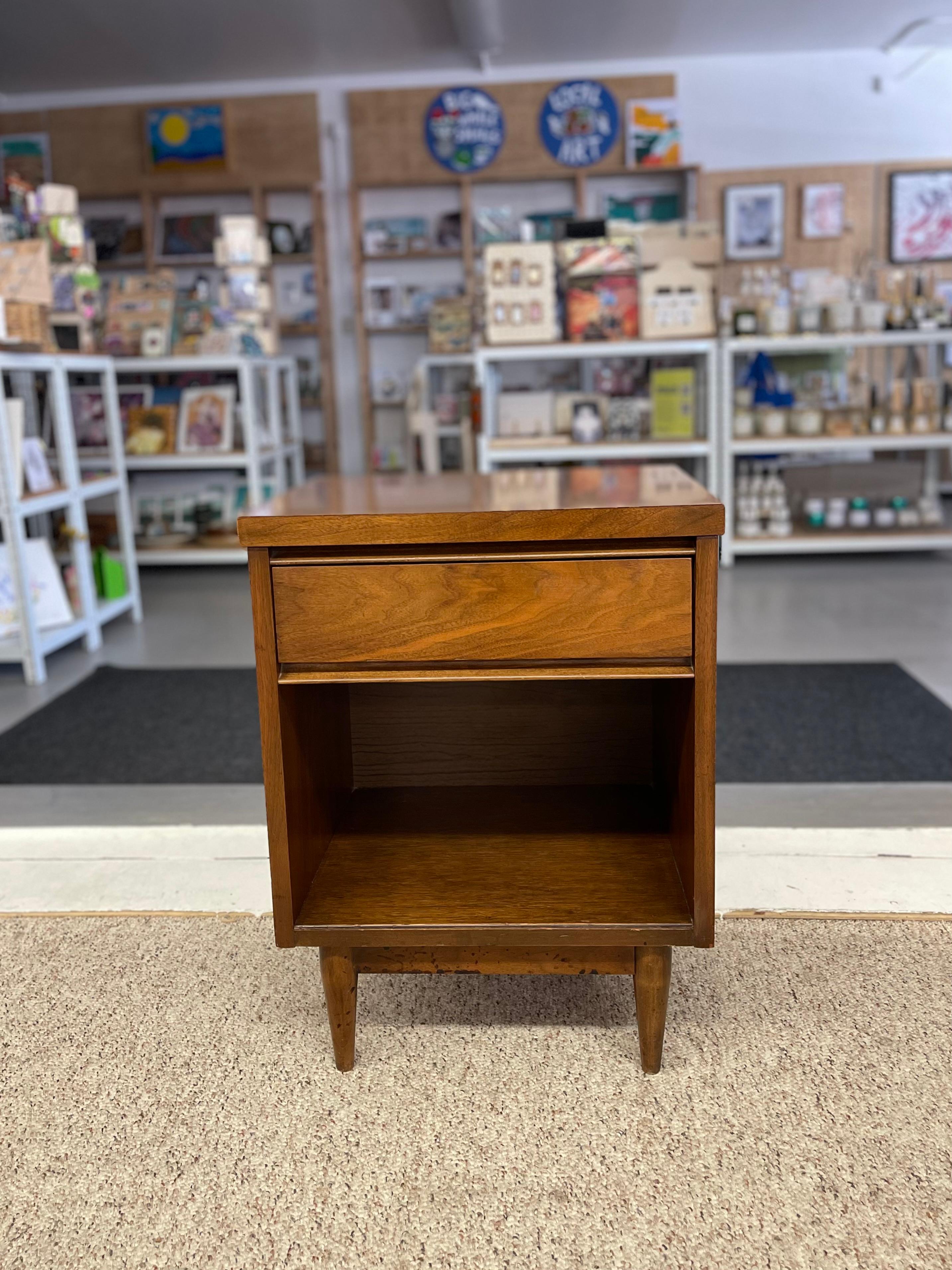  This vintage mid-century modern nightstand showcases clean lines, practical design, and rich walnut wood grain typical of 1950s American craftsmanship. The piece features a single flush-mounted drawer with a minimalist integrated lip pull, sitting