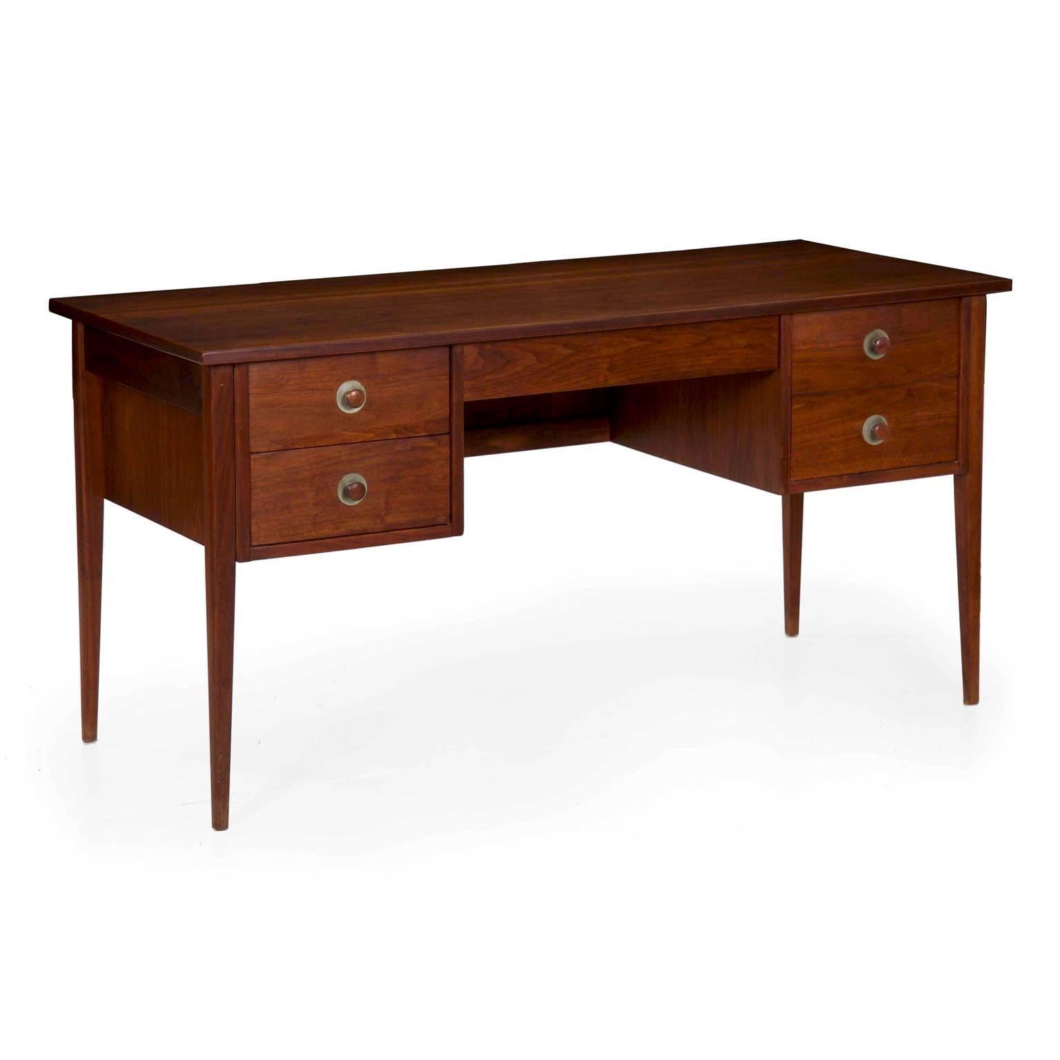 This is an attractive 1960s vintage writing desk crafted of walnut and walnut veneers over oak secondaries in the machine-dovetailed drawers. It is sleek and angular with nice turned handles backed by steel accents, the pair of stacked drawers on
