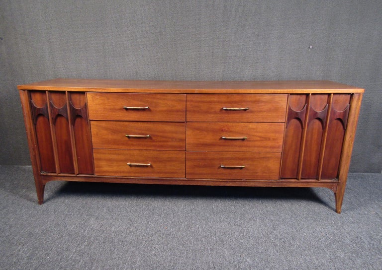 This Mid-Century Modern server by Kent Coffey features rich walnut woodgrain, brass and wooden accents, and abundant storage with twelve drawers. The server has six center drawers with brass handles, along with three smaller drawers on each end