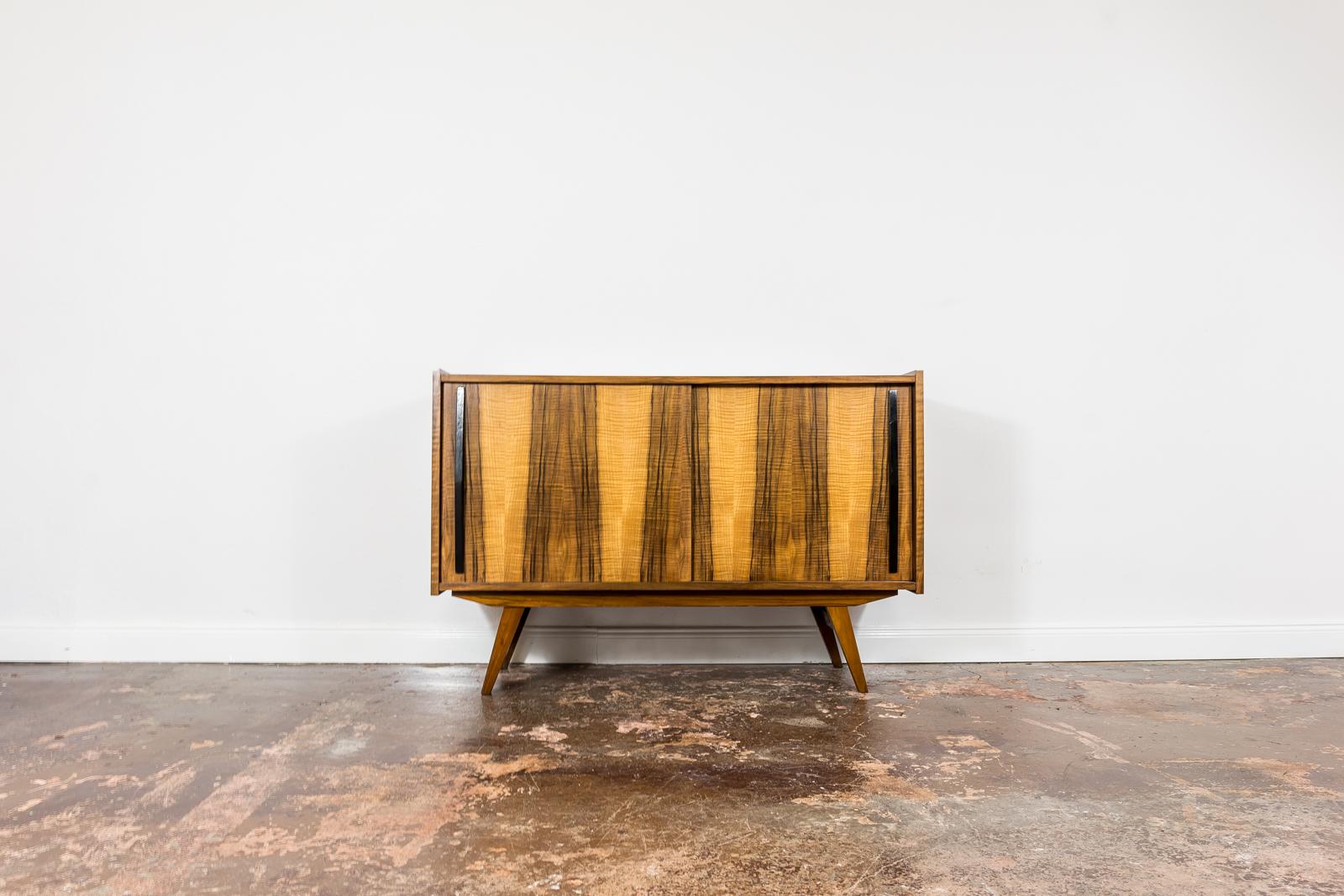 Walnut veneer sideboard from Słupskie Furniture Factory 1960's, Poland.
Sideboard has 2 sliding doors with black handles, and removable shelf.
Case raised on solid wooden construction and legs.
This item has been restored and refinished enhancing