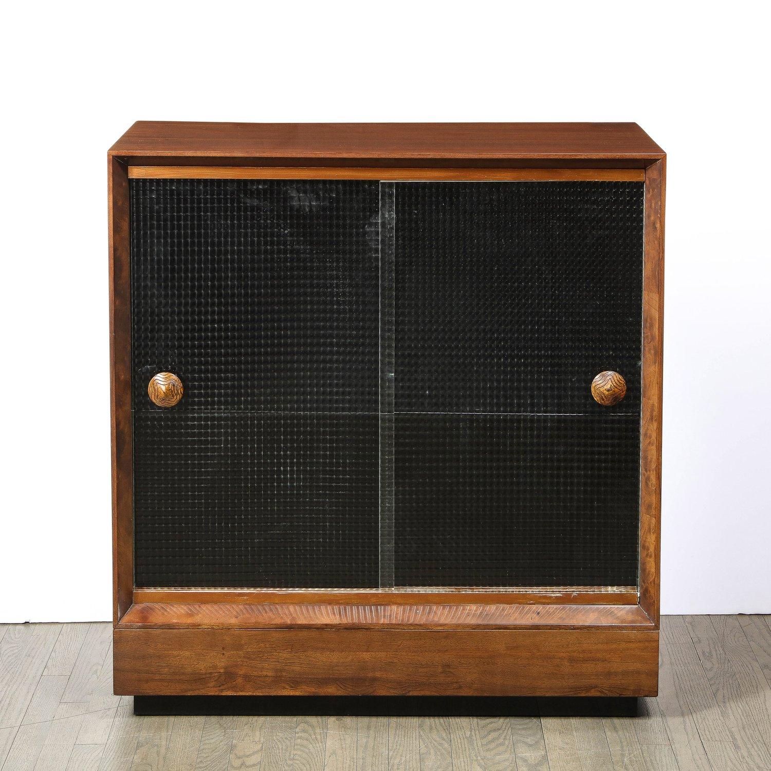 This beautiful Mid-Century Modern dry bar/ cabinet was realized by the illustrious American designer Gilbert Rohde, circa 1943. It features a volumetric rectangular body in bookmatched acacia with two sliding doors and a black lacquer inset base.