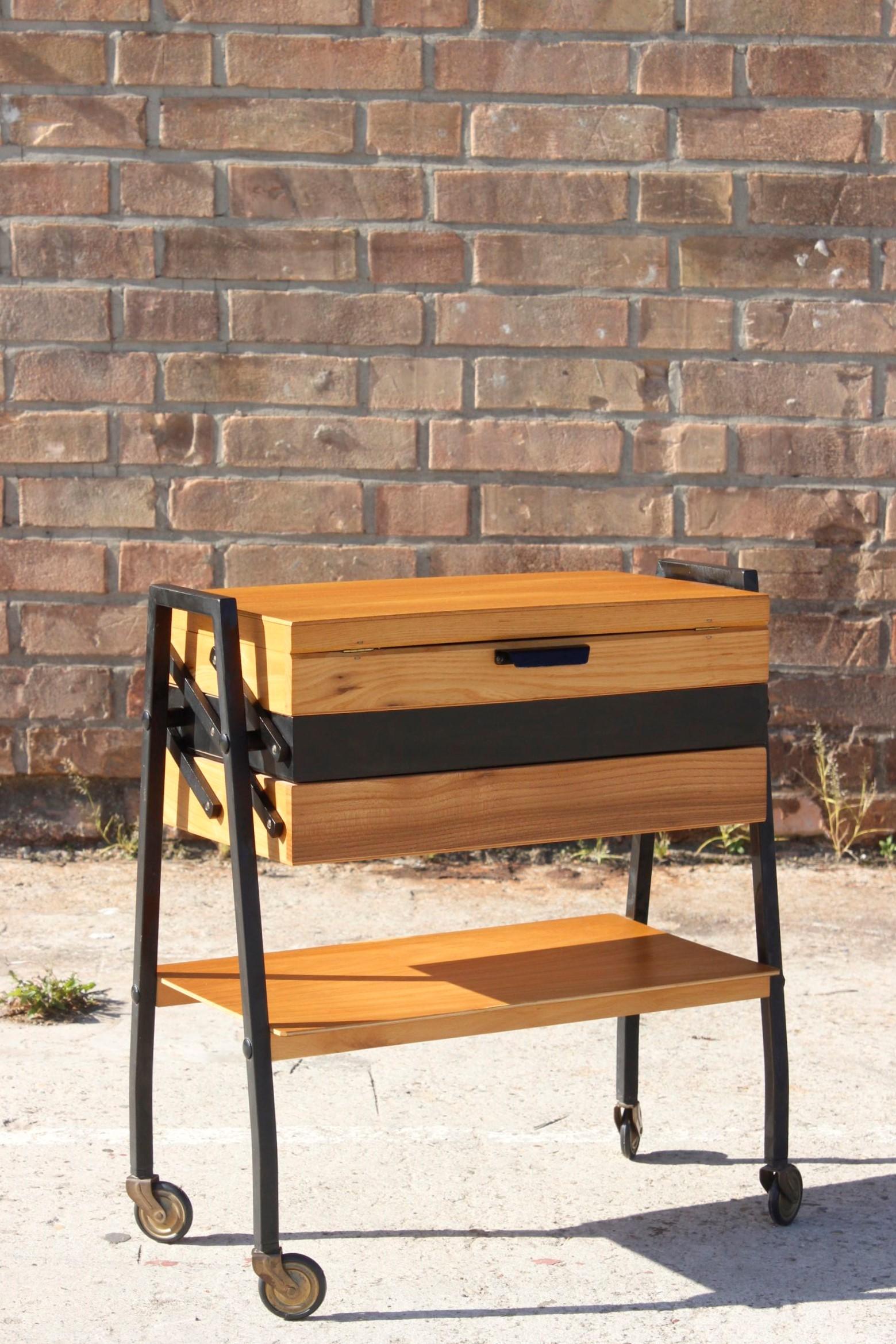 Mid-century modern sewing table on wheels, made out of light varnished wood and steel painted black. 

Unfolds into several compartments for organizing sewing equipment, also has a simple drawer. 

Can be used for its original purpose or as a