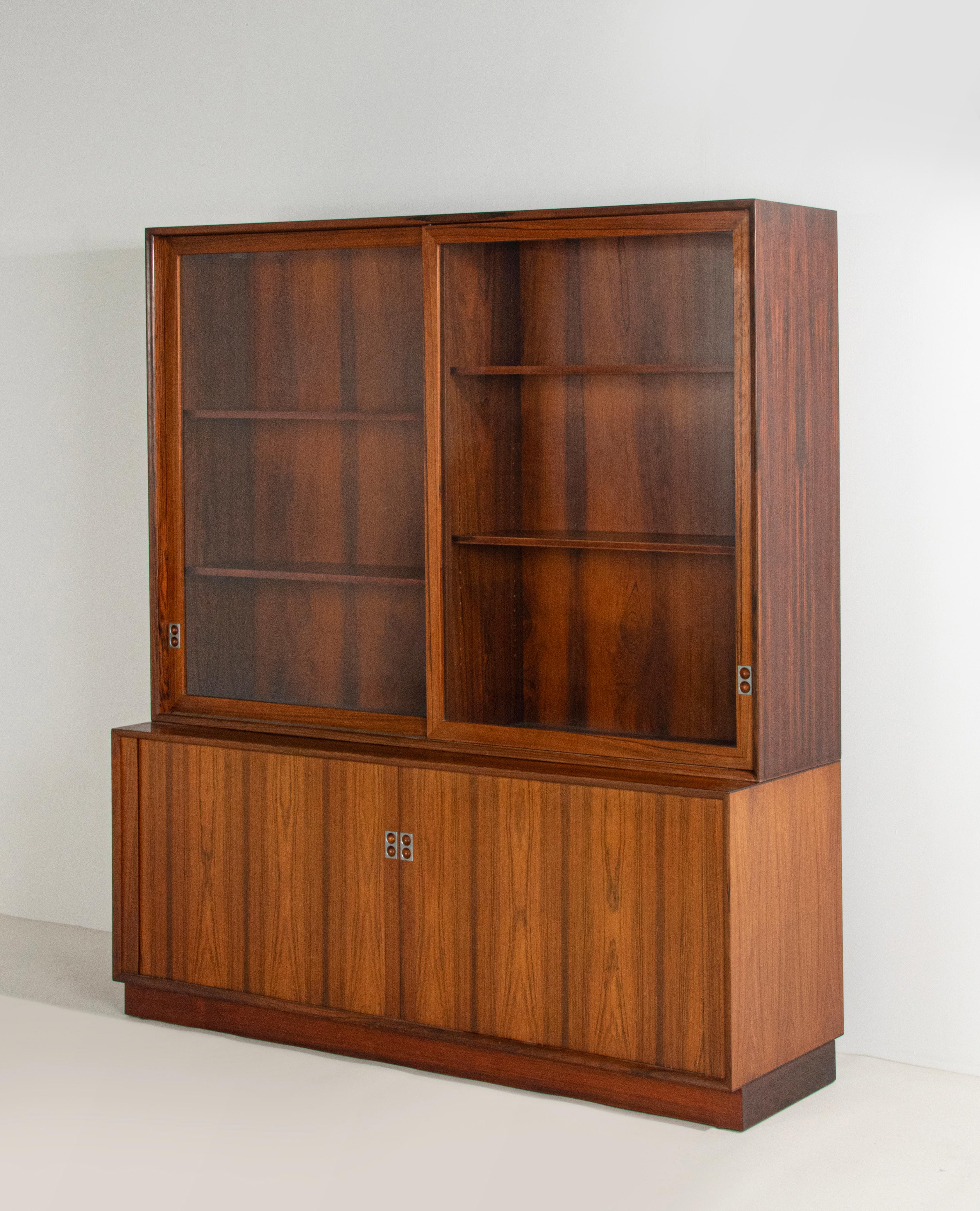 A Mid-Century Modern veneered bookcase with spacious interior. Sliding glass doors with metal handles. The base part has two tambour sliding doors, with two extra drawers. The design is by Arne Vodder, made by Sibast Møbler. All doors opens