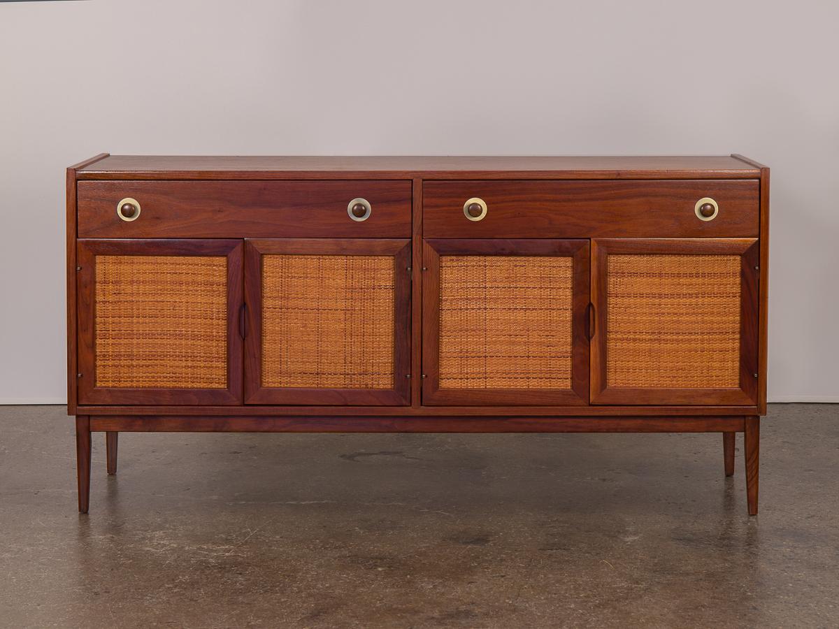 A classic Mid-Century Modern walnut credenza. Features two top drawers with circular pulls and pretty brass medallion hardware. Additional woven cane cabinets with swing doors, each with an adjustable shelf. Credenza is in excellent vintage