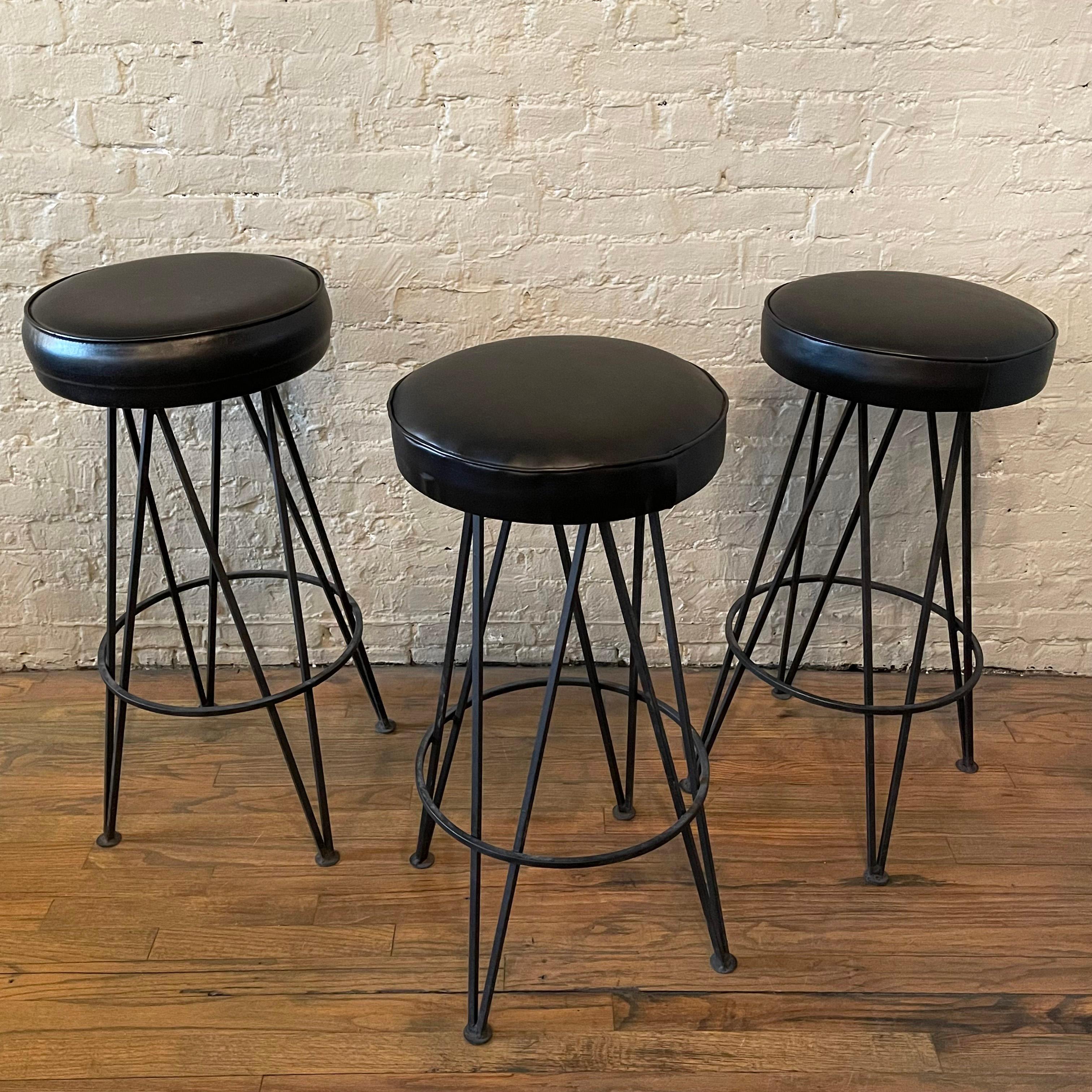 Set of three, classic, mid century modern bar stools feature wrought iron hairpin bases with 14 inch round black vinyl seats. The 14 inch diameter footrests are at 10 inches height. The seats do not swivel and one of the stools is slightly taller at