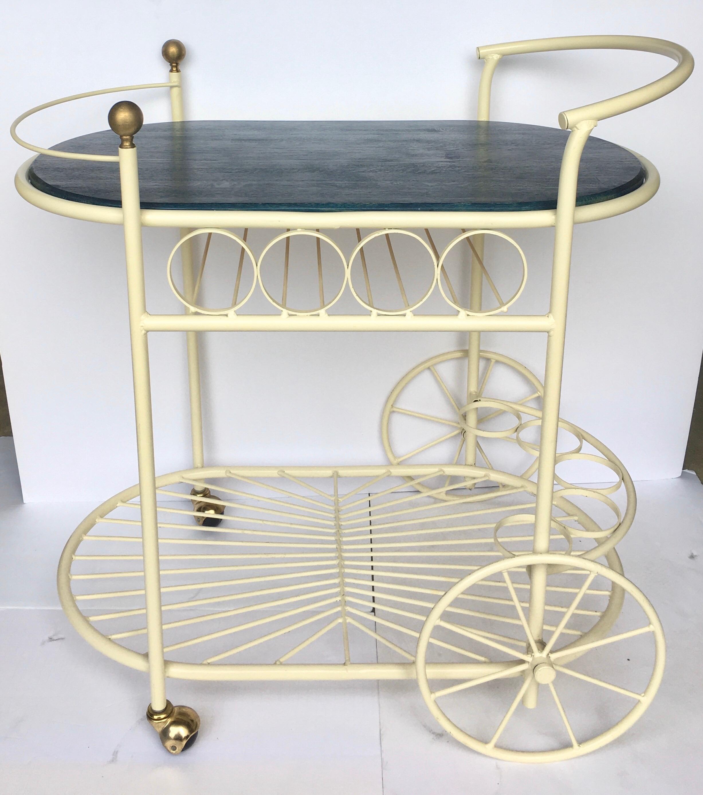 Charming Mid-Century Modern cream painted wrought iron bar cart or drinks trolley with removable navy blue stained wood top. Design features include two oval shaped tiers, nine wine bottle holders, and two decorative brass finials.