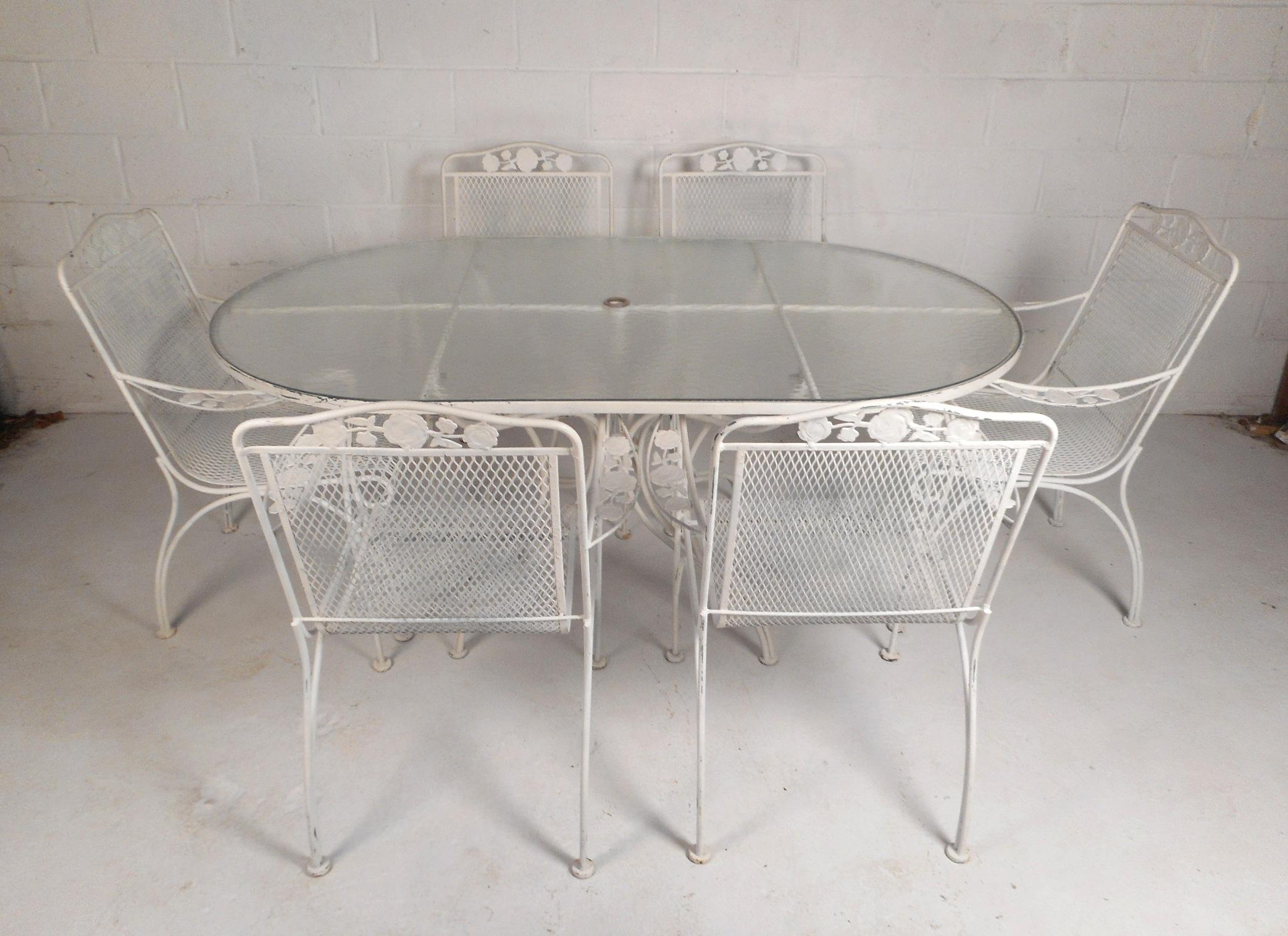 This beautiful vintage modern patio set includes a large oval dining table with a tempered frosted glass top and six sculpted dining chairs. The sleek set of chairs feature a bent rod iron frame, floral detail, and grated seats offering plenty of