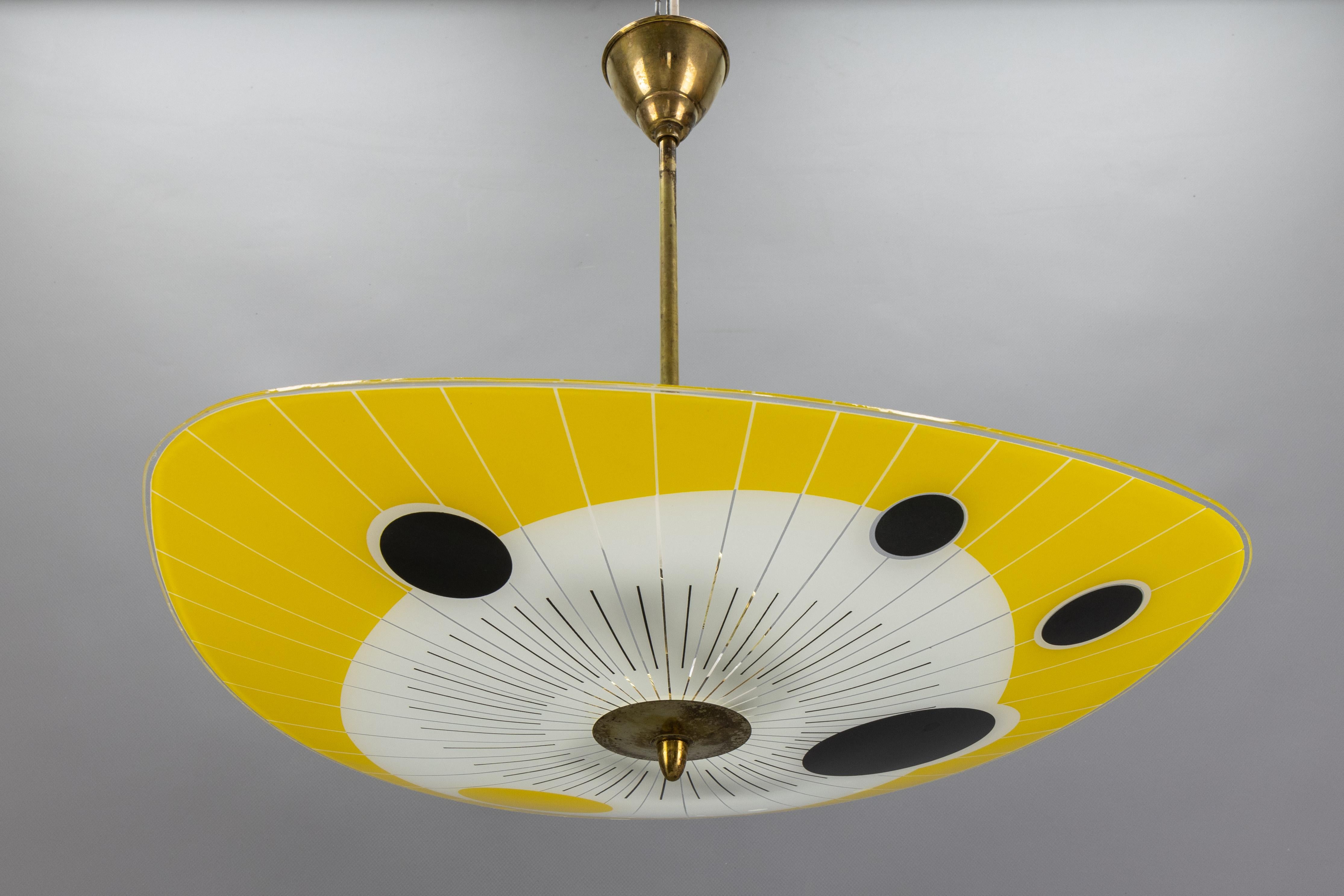 Mid-Century Modern yellow, black, and white glass and brass two-light pendant light, France, 1950s. 

This striking ceiling fixture features a softly curved, triangular glass shade adorned with vibrant yellow segments and bold circular motifs in