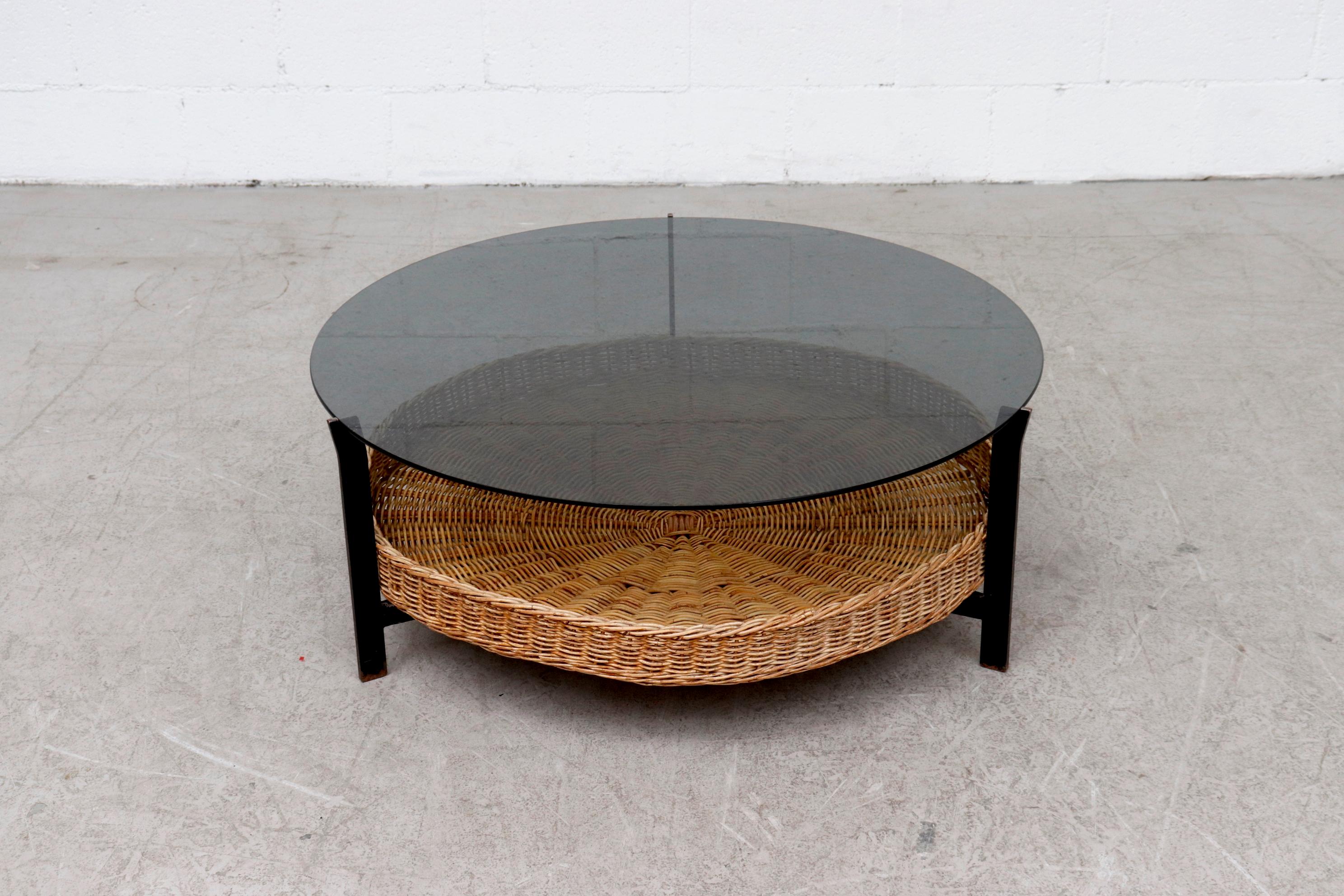 Mid-century smoked glass coffee table with lower woven wicker basket on black enameled metal frame. In original condition with heavy patina to the frame and glass. Visible scratching to the glass. Wear is consistent with its age and use. Others