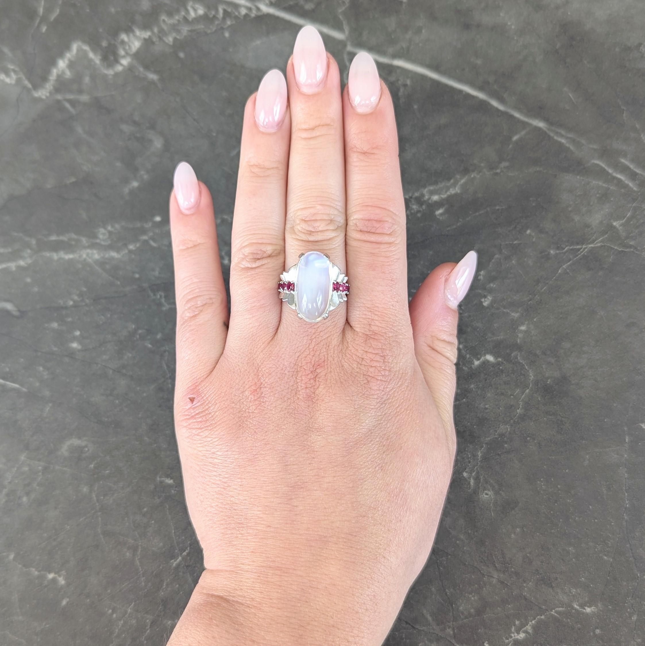 Centering an oval shaped moonstone cabochon measuring 20.0 x 10.0 mm
Translucent colorless body displaying vivid blue adularescence
Prong set in basket and flanked by fanning stepped shoulders
Prong set with graduated round cut rubies
Weighing
